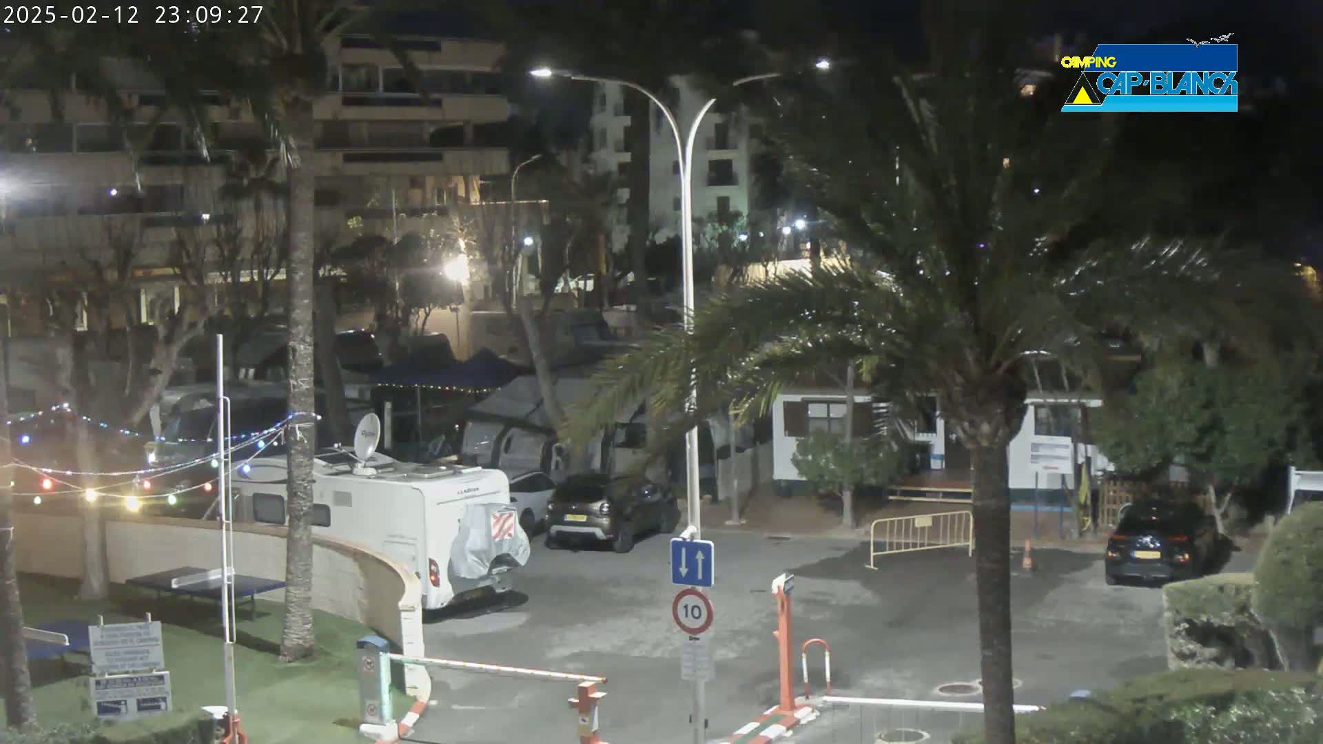 A nighttime scene in a campground shows several RVs and cars parked among palm trees and buildings, with string lights strung along a fence.