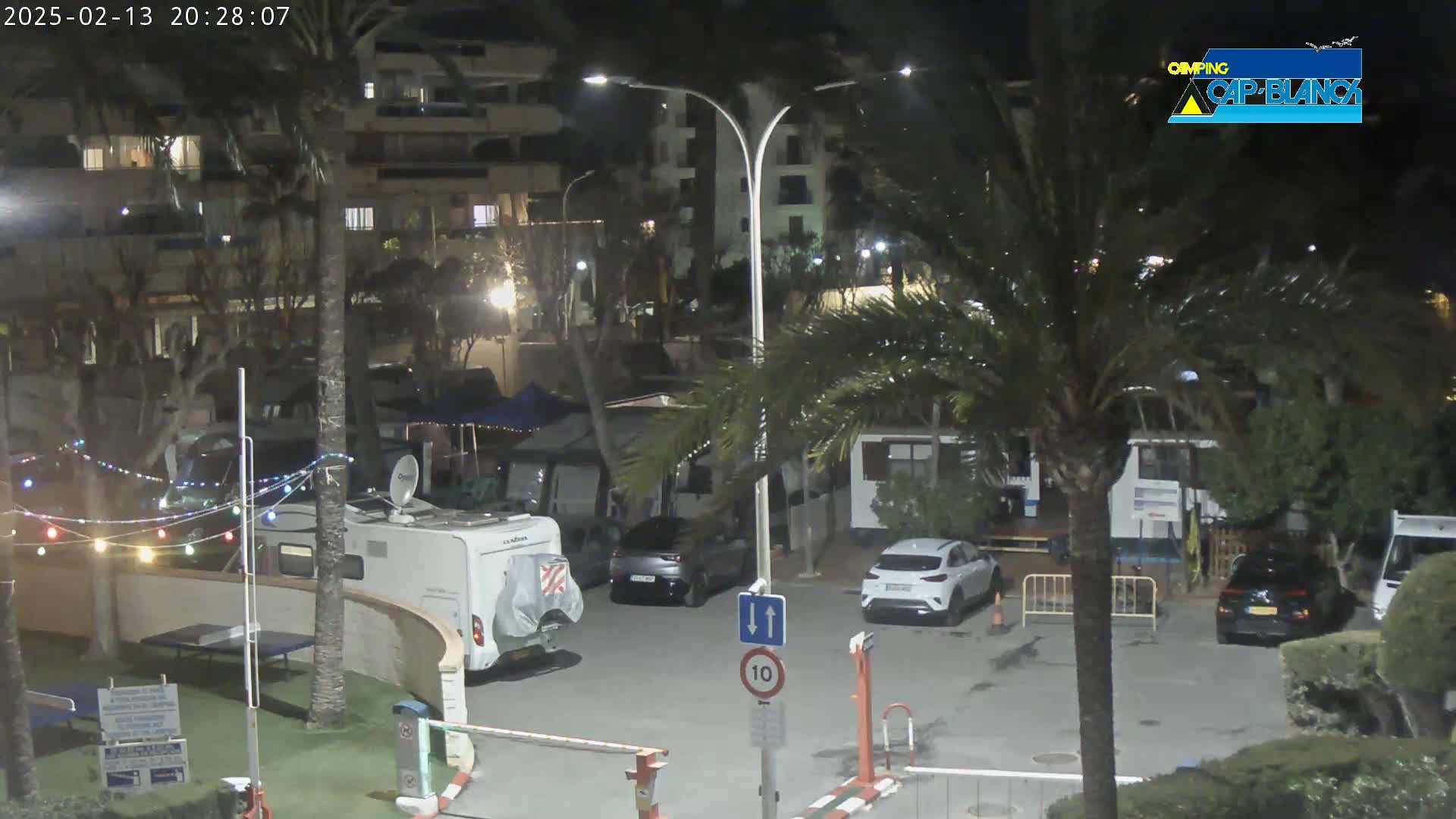 At night, a parking lot with several cars and RVs is illuminated by streetlights, surrounded by palm trees and buildings.