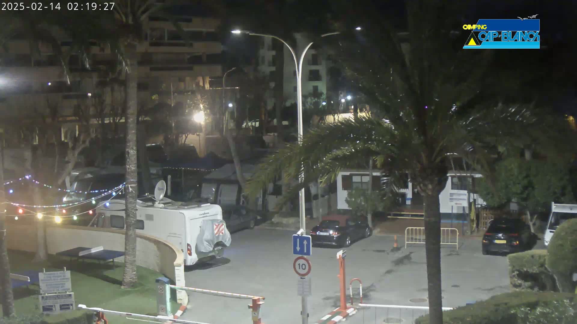 A nighttime scene shows a parking lot with several vehicles, including RVs and cars, next to buildings and palm trees under dark, clear skies.