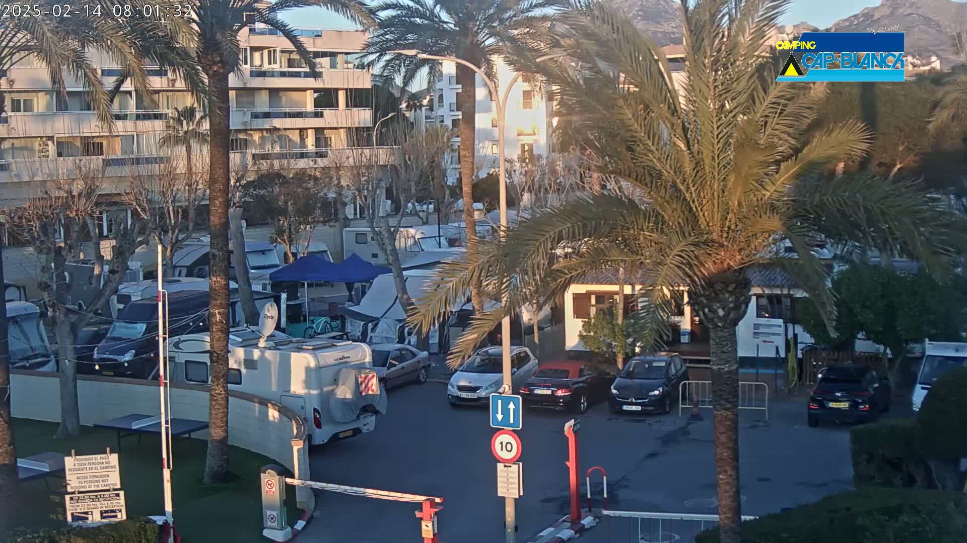 A parking lot with numerous RVs and cars is situated near a multi-story building under a partly sunny sky.