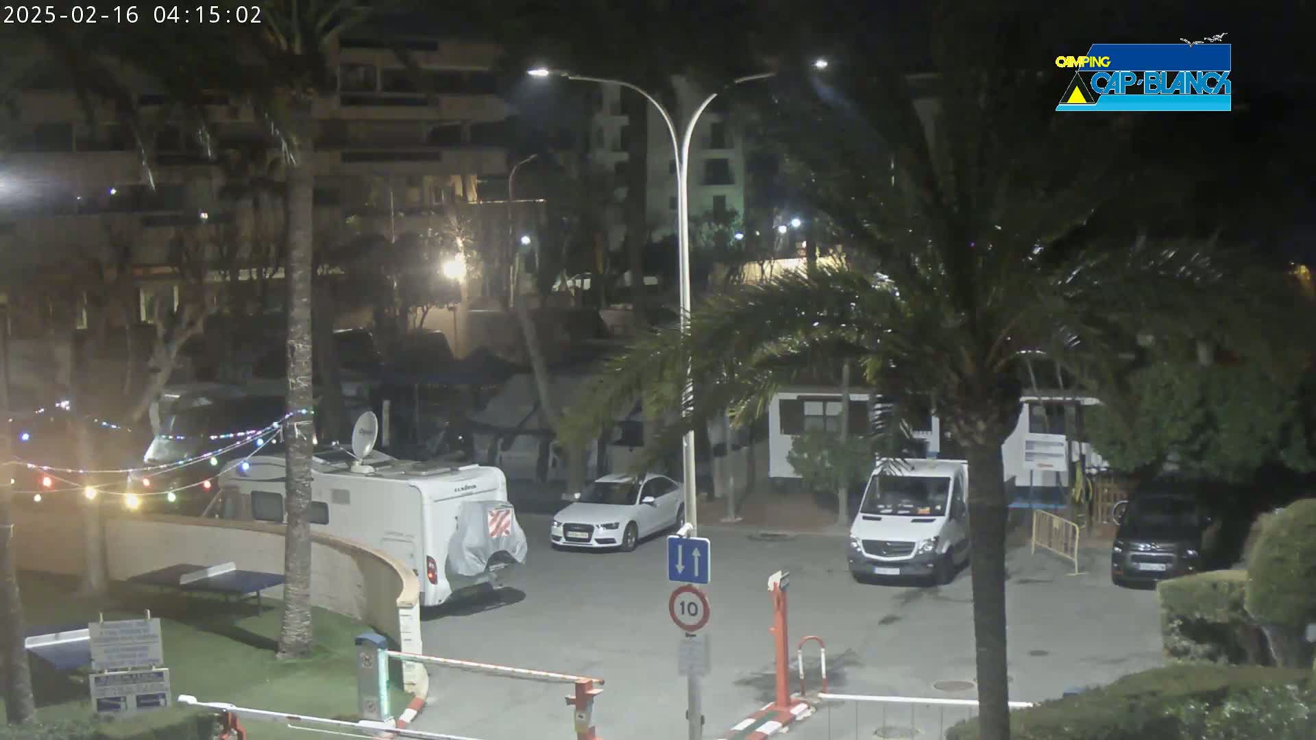 A nighttime view of a parking lot with several vehicles, including a camper van and a car, situated near palm trees and apartment buildings under low-light conditions.