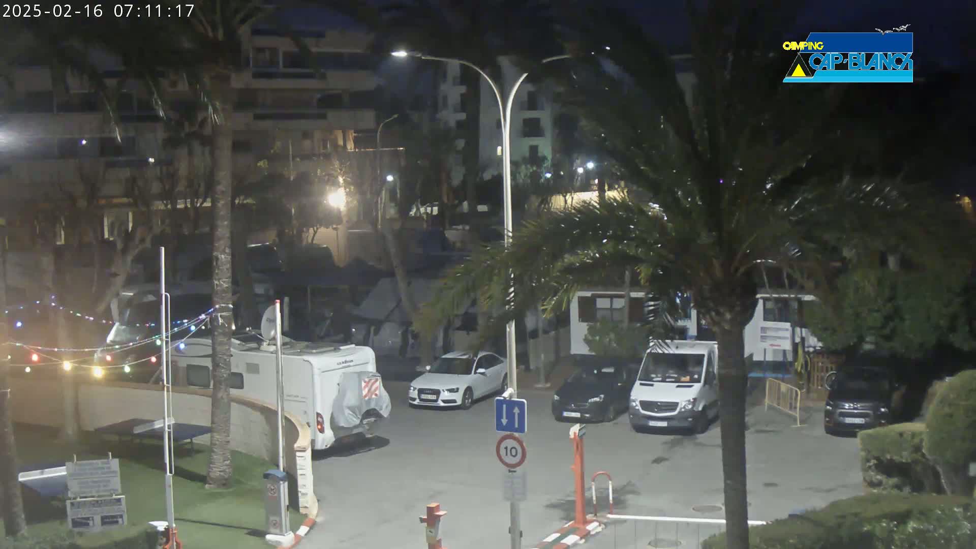 At night, under a dark sky, a parking lot contains several cars, camper vans, and palm trees, with string lights strung between some of the vehicles.