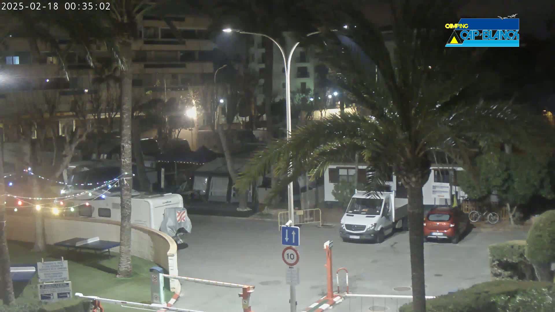 A nighttime view of a parking lot with RVs and cars, illuminated by streetlights, surrounded by palm trees and buildings.