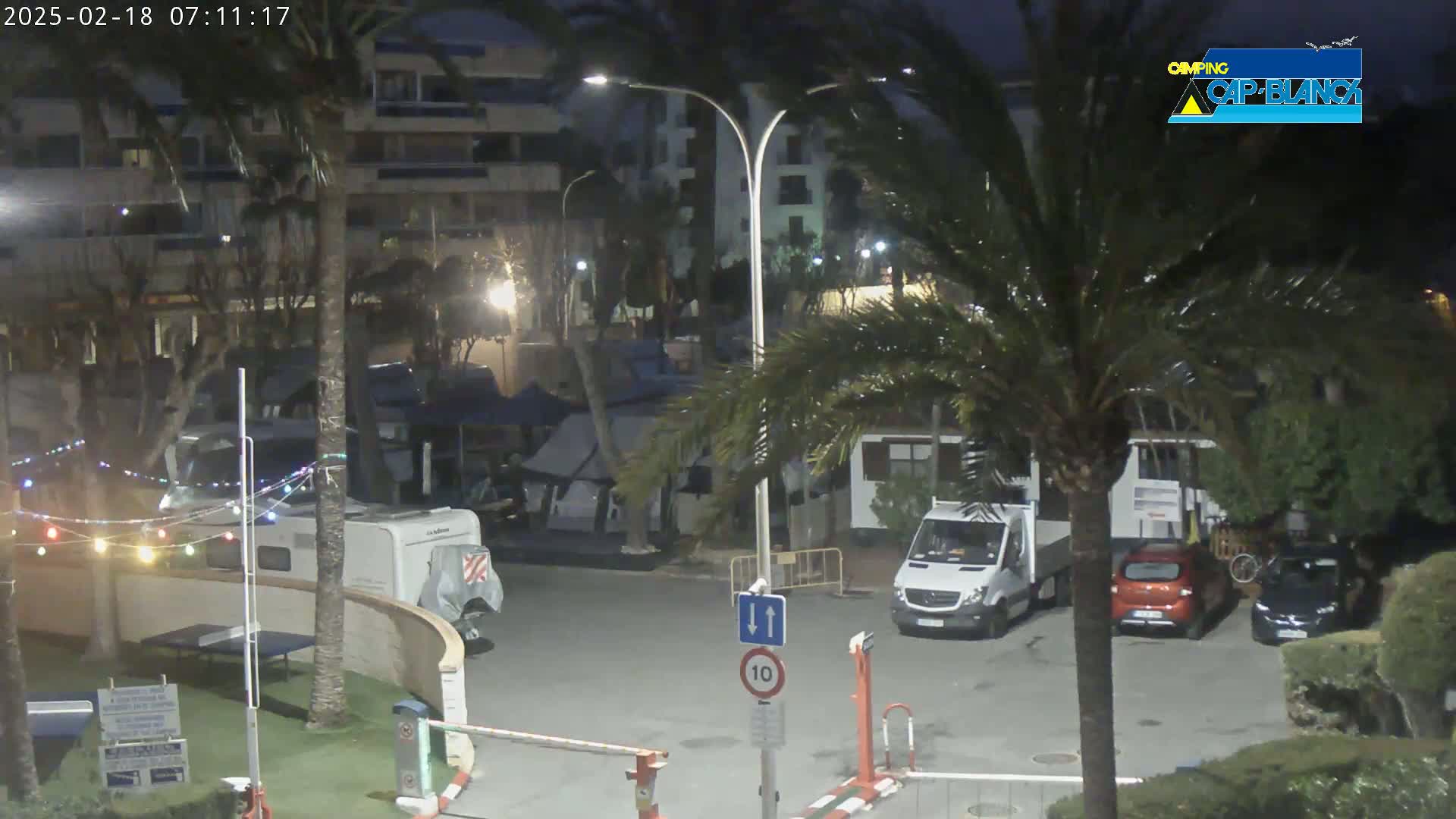 A nighttime view of a parking lot with several vehicles, including a camper van, surrounded by palm trees and buildings, under partly cloudy skies.