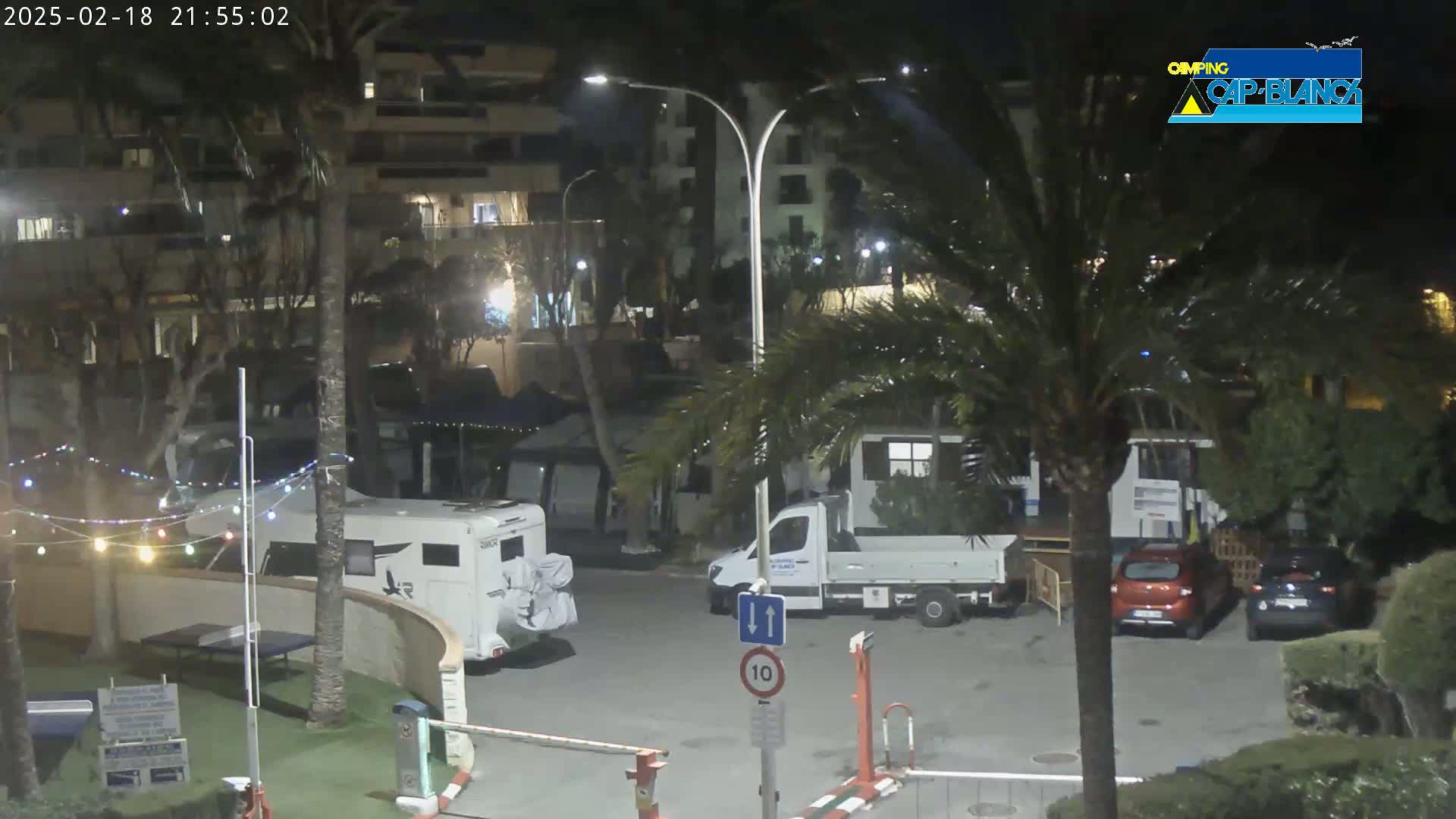 At night, a parking lot with several vehicles, including camper vans and cars, is illuminated by streetlights amidst palm trees and buildings.