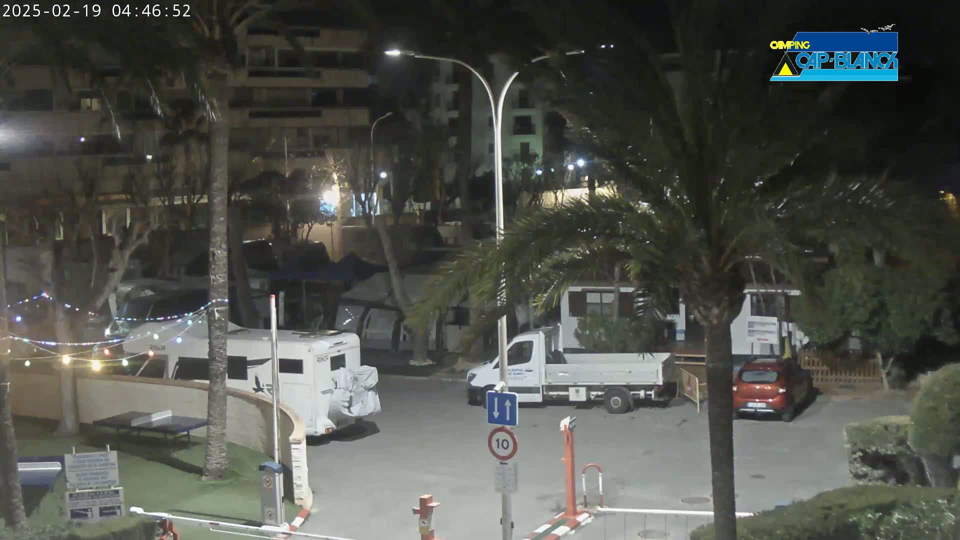 At night, a parking lot with several vehicles, including a campervan and a pickup truck, is illuminated by streetlights amidst palm trees and buildings.