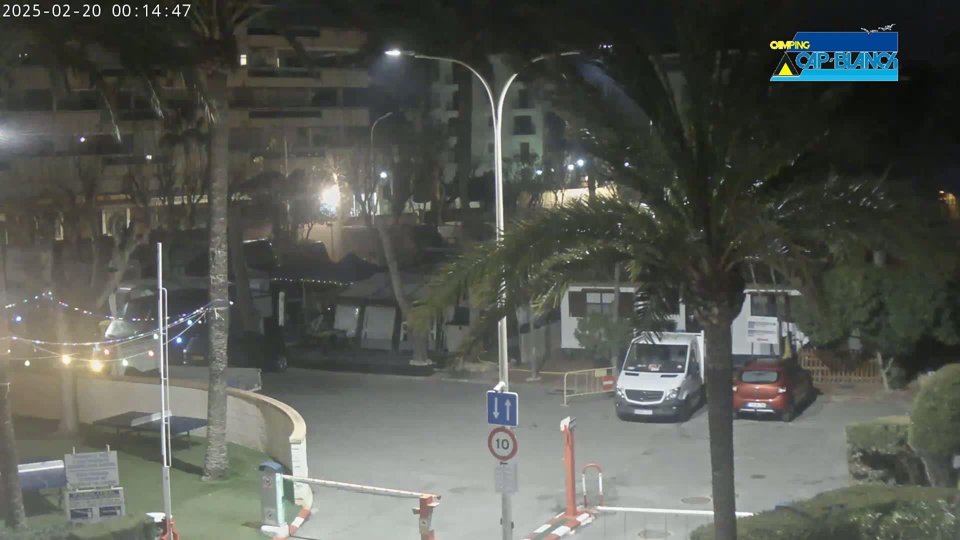 A nighttime outdoor scene shows a parking lot with a van and car, surrounded by palm trees, buildings, and string lights.