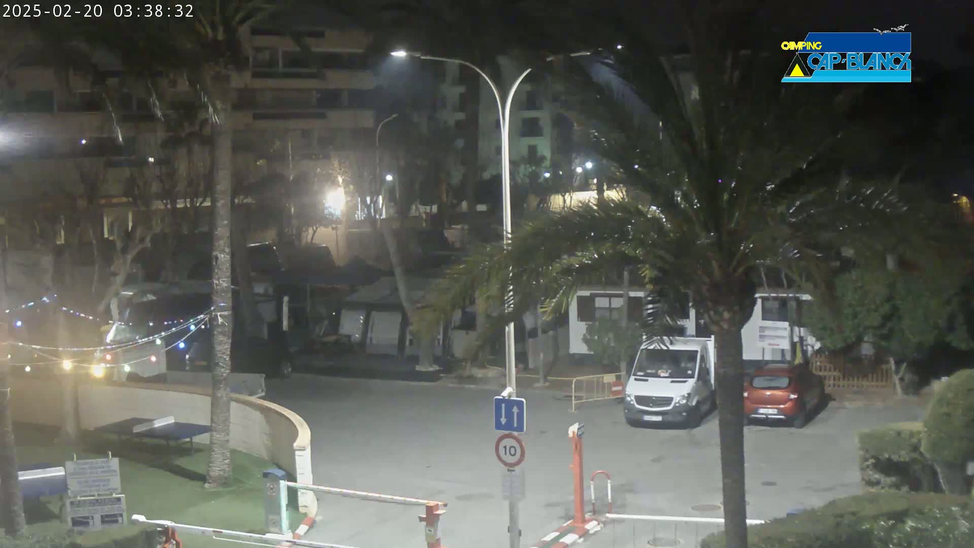 A nighttime view of a parking lot and surrounding buildings under a dark, possibly rainy sky, with palm trees and string lights visible.