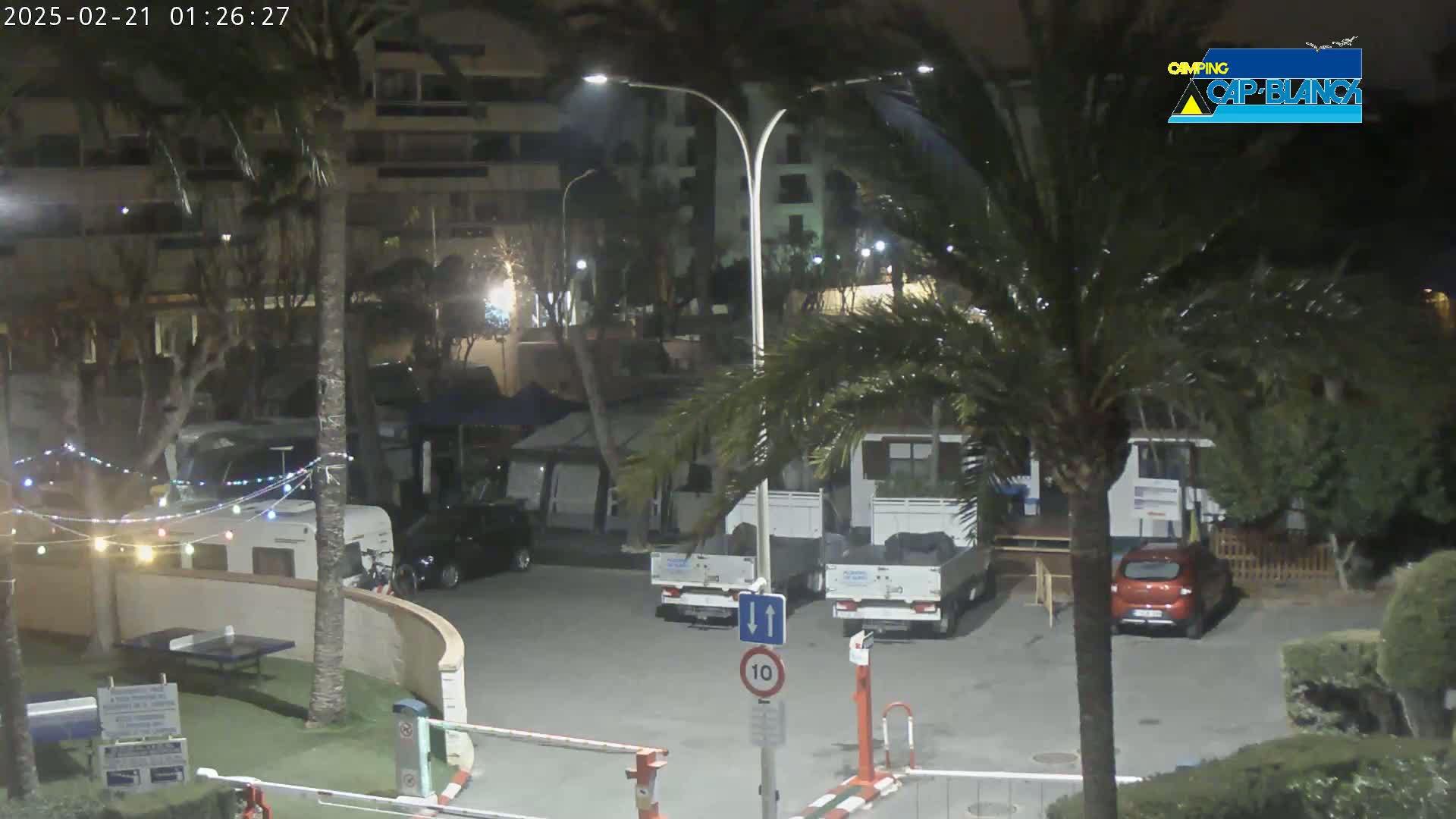 A nighttime view of a parking lot with cars, RVs, and palm trees, under partly cloudy skies.
