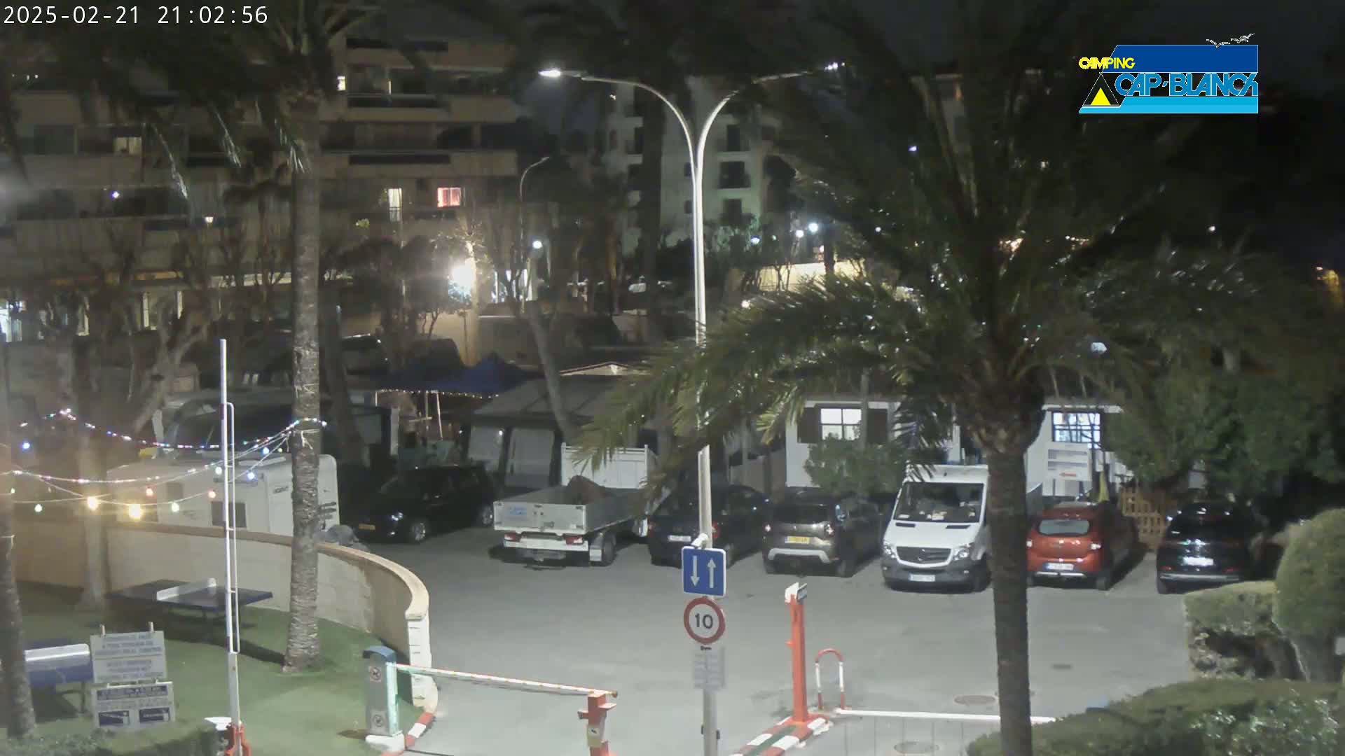A nighttime view of a parking lot with several vehicles, palm trees, and buildings under partly cloudy skies.