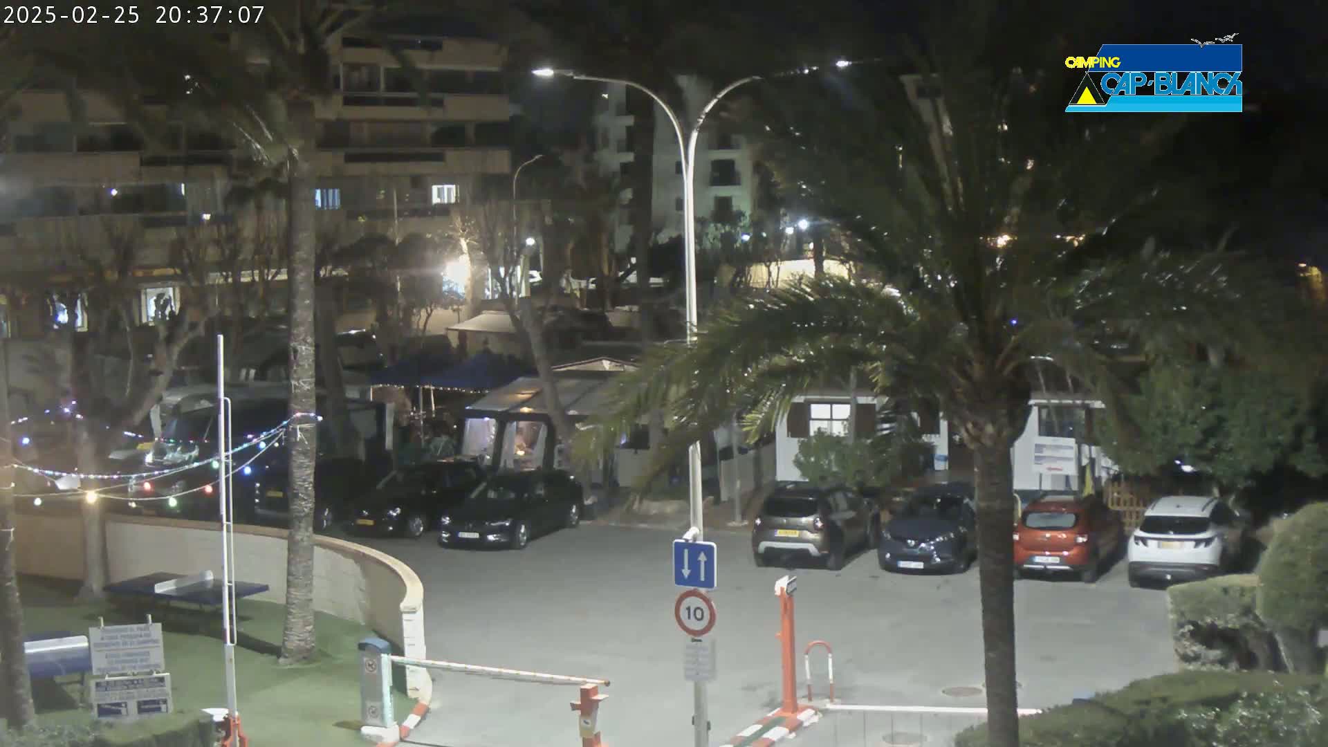 A nighttime parking lot scene with several cars parked near buildings and palm trees under partly cloudy skies.