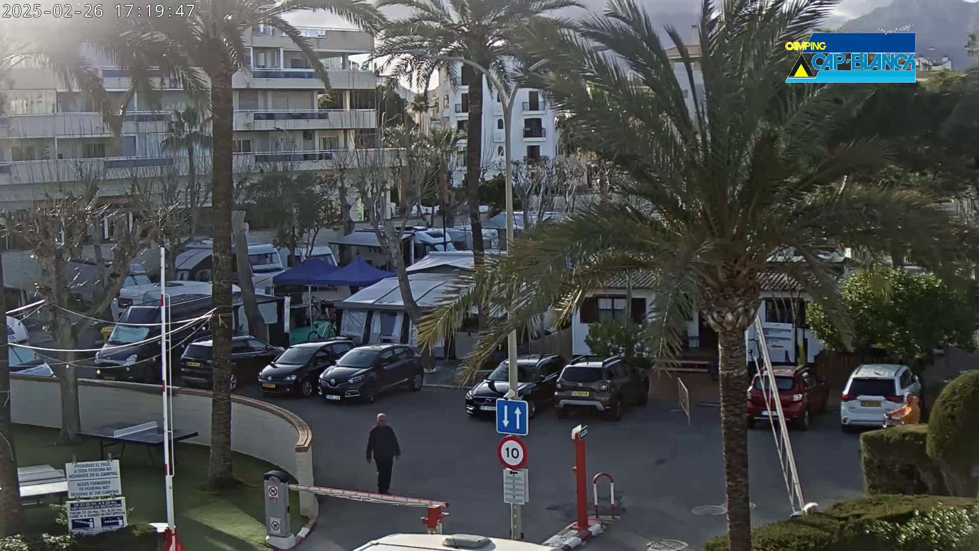 A parking lot with numerous cars and campervans is situated near apartment buildings and palm trees under partly cloudy skies.