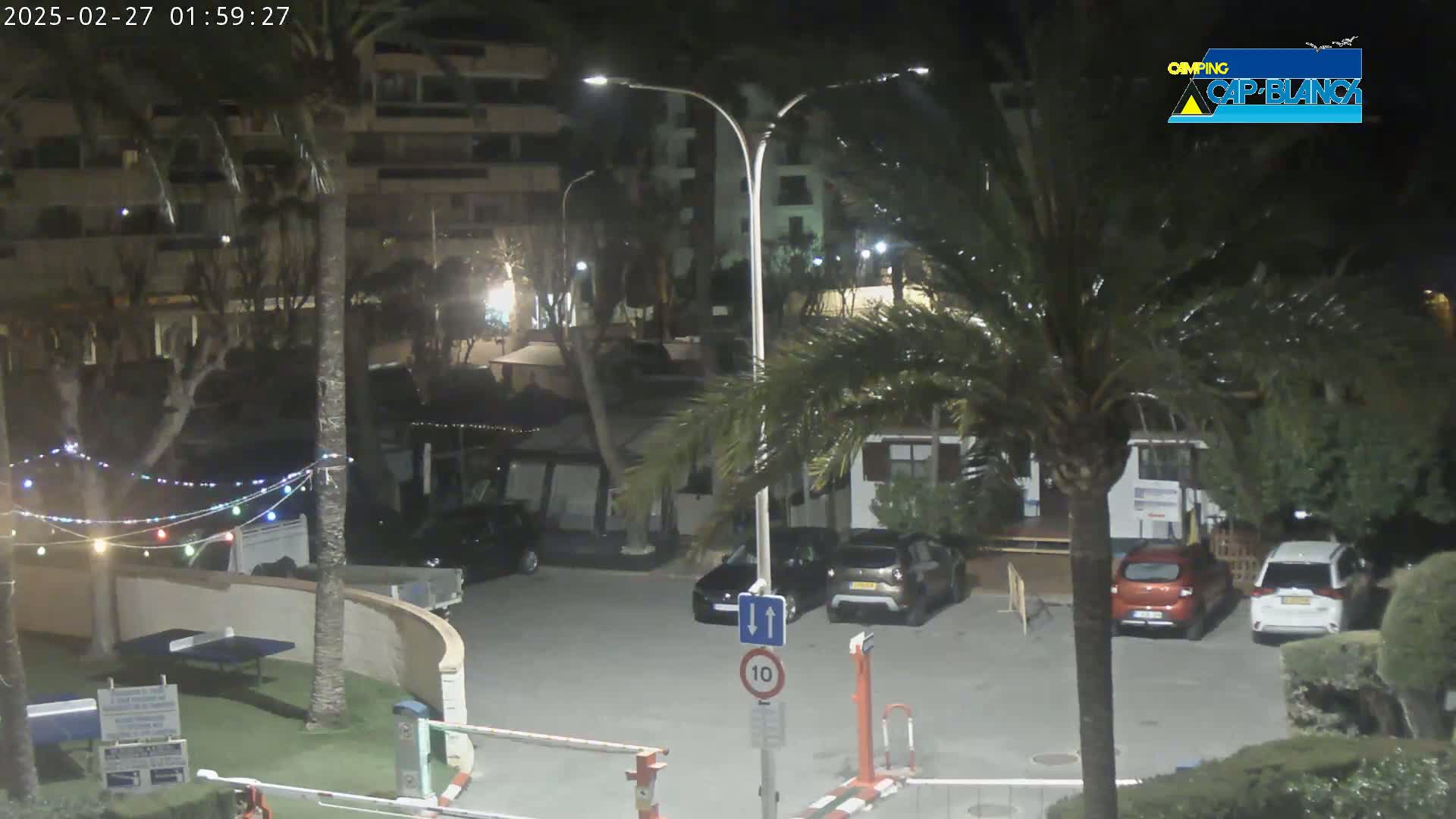 A nighttime view of a parking lot with several cars, palm trees, and a building, under partly cloudy skies.