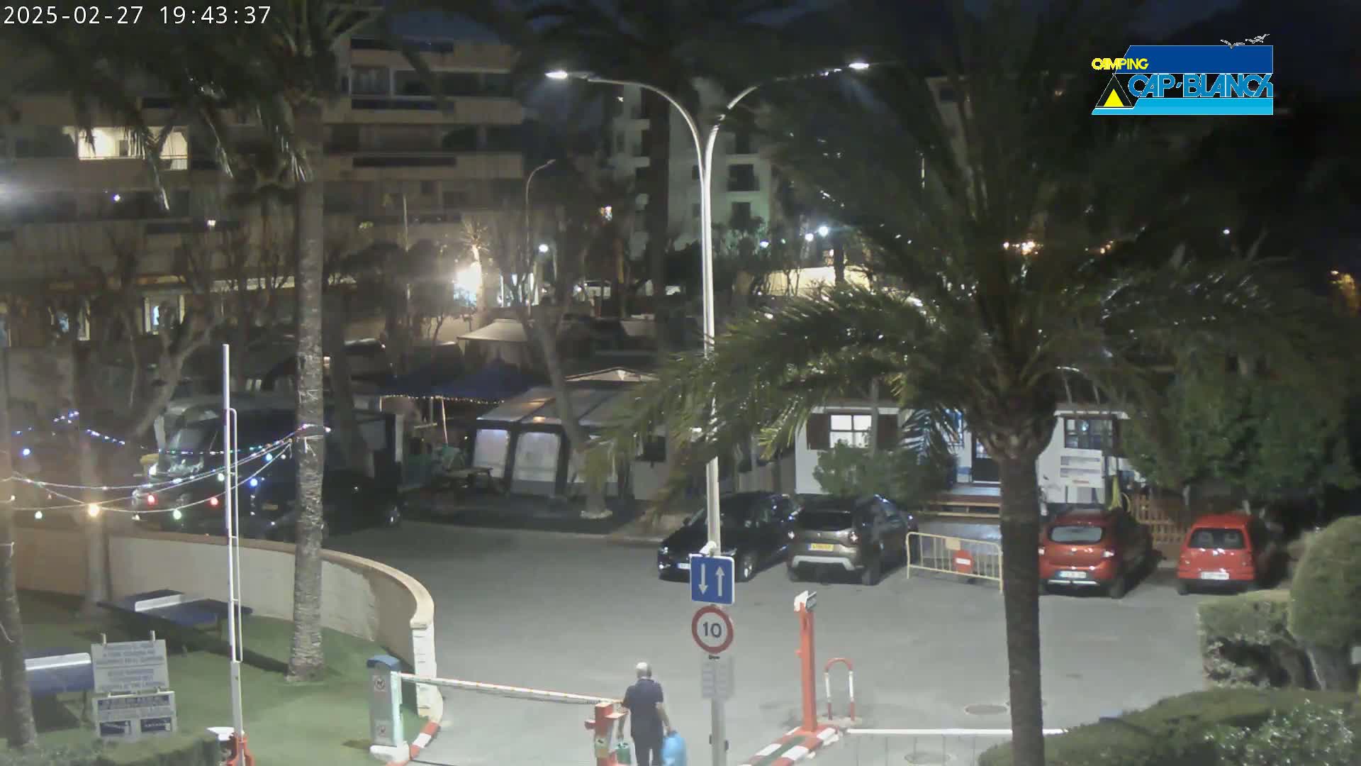 At night, under partly cloudy skies, a parking lot is partially visible, surrounded by palm trees and buildings, with a few cars parked and a person walking.