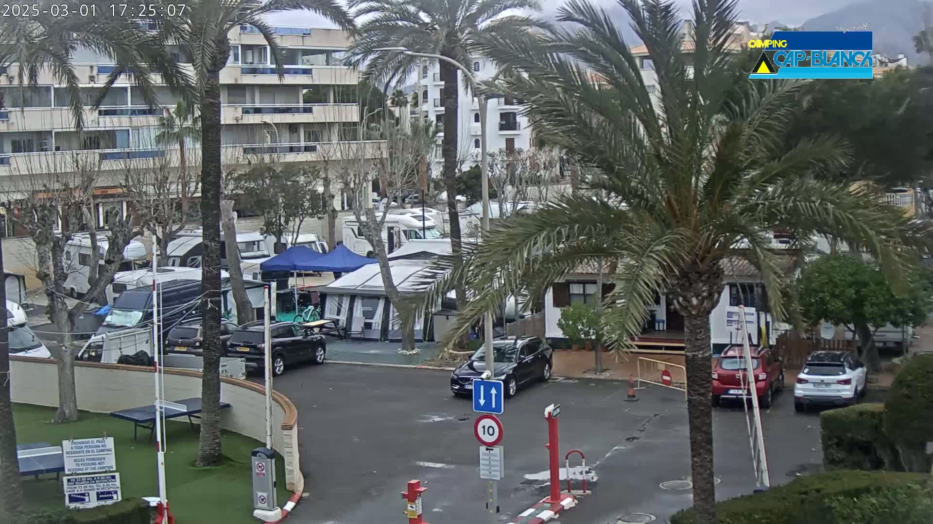 A parking lot with numerous RVs and tents is situated next to a multi-story building under an overcast sky.