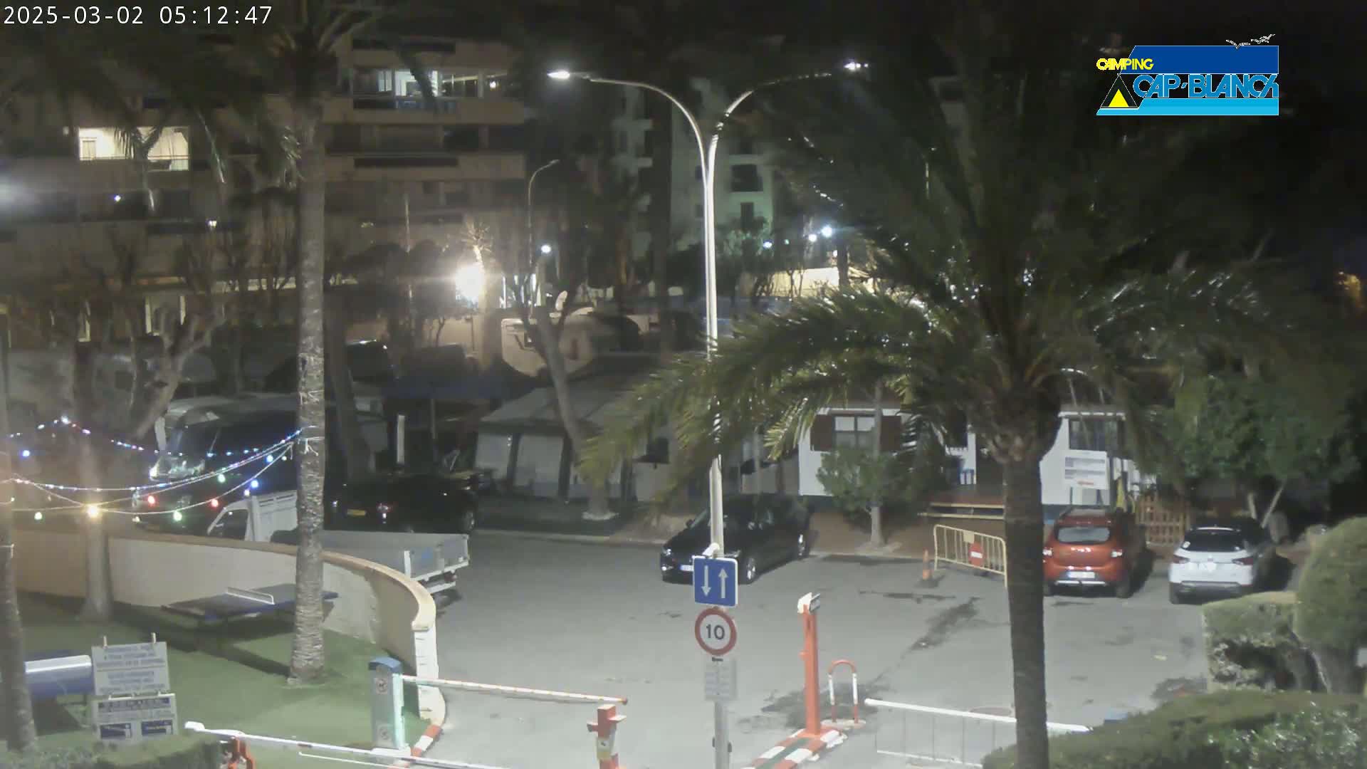 A nighttime view of a parking lot with several cars, surrounded by palm trees and buildings, under partly cloudy skies.
