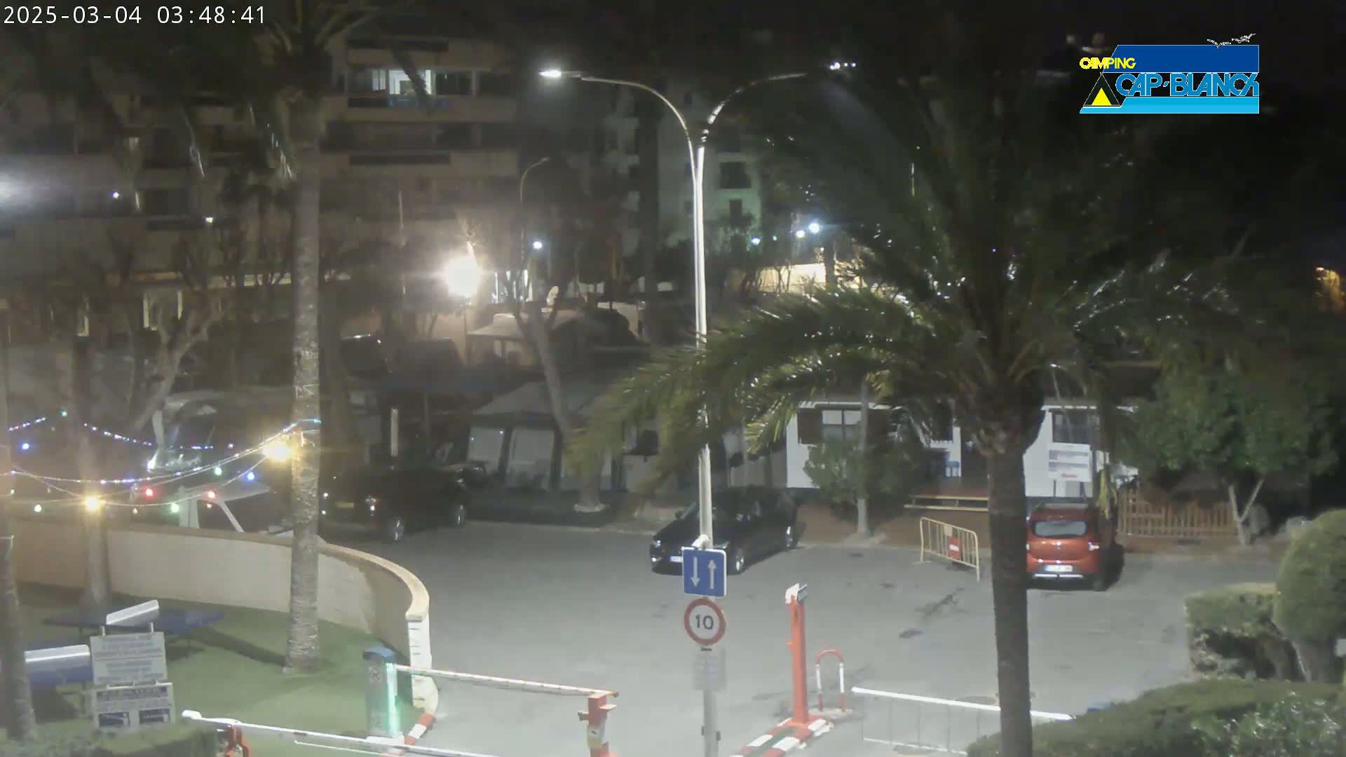 A nighttime view of a parking lot with several cars, palm trees, and buildings under slightly rainy conditions.