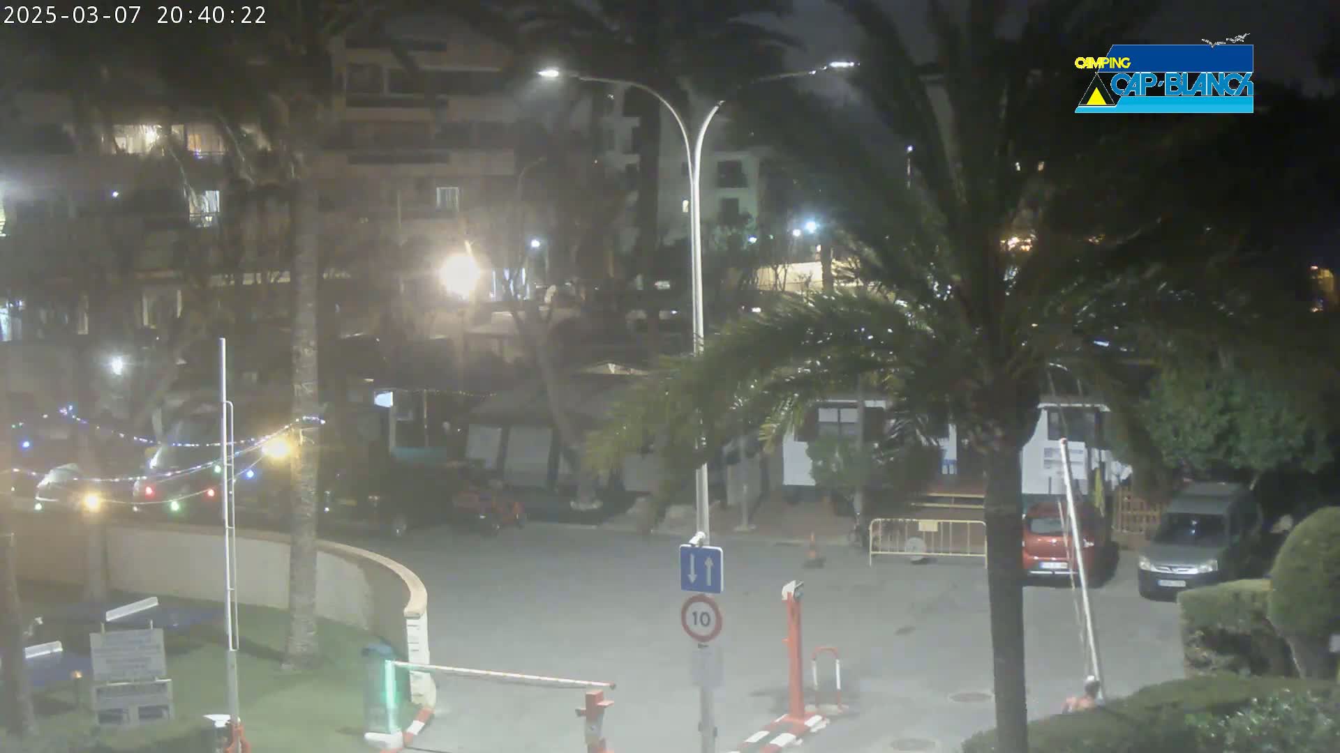 A nighttime street scene shows a partly cloudy sky, palm trees, streetlights, and several cars parked near buildings with string lights strung between them.