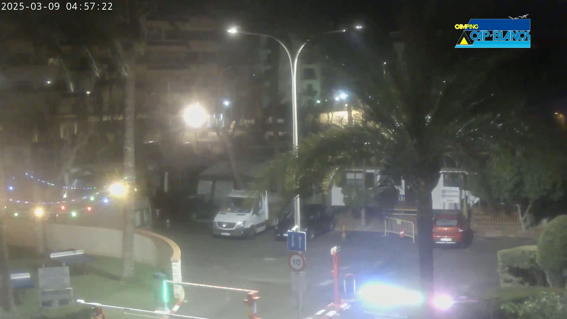 A nighttime view of a street scene with parked cars and a van, illuminated by streetlights and string lights, under overcast conditions.