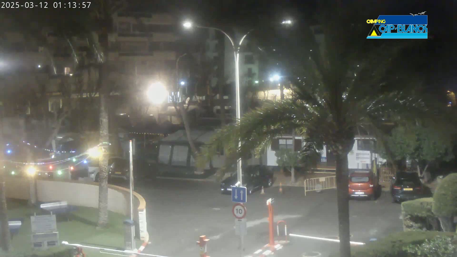 A nighttime view of a parking lot with several cars, palm trees, and string lights, under partly cloudy skies.