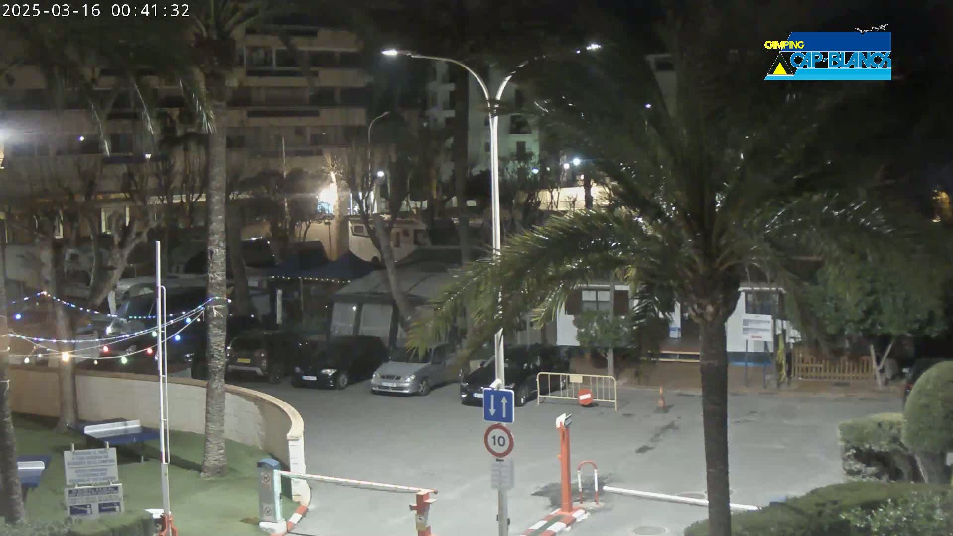 A nighttime view of a parking lot surrounded by palm trees and buildings, with a few cars parked and string lights visible.