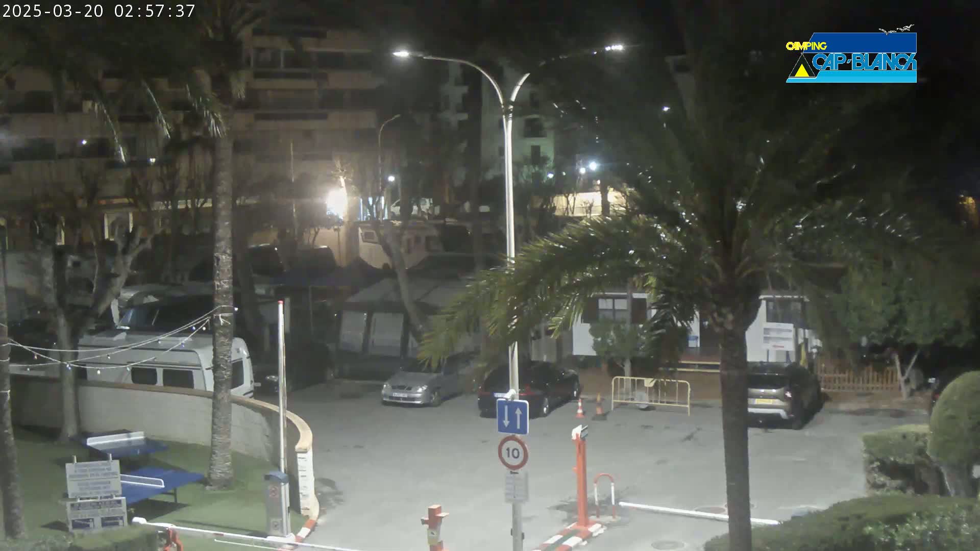 A nighttime view of a parking lot with several cars and RVs, surrounded by palm trees and buildings under partly cloudy skies.