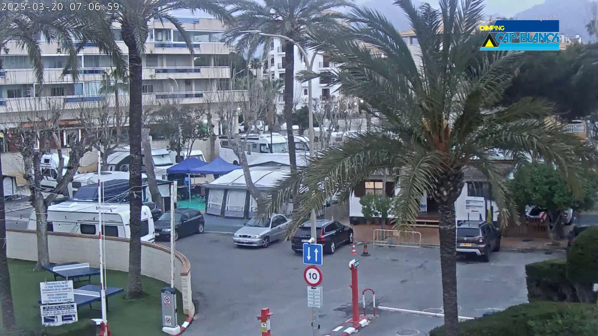 A cloudy day overlooking a campground with numerous RVs and tents, situated next to a multi-story building and palm trees.