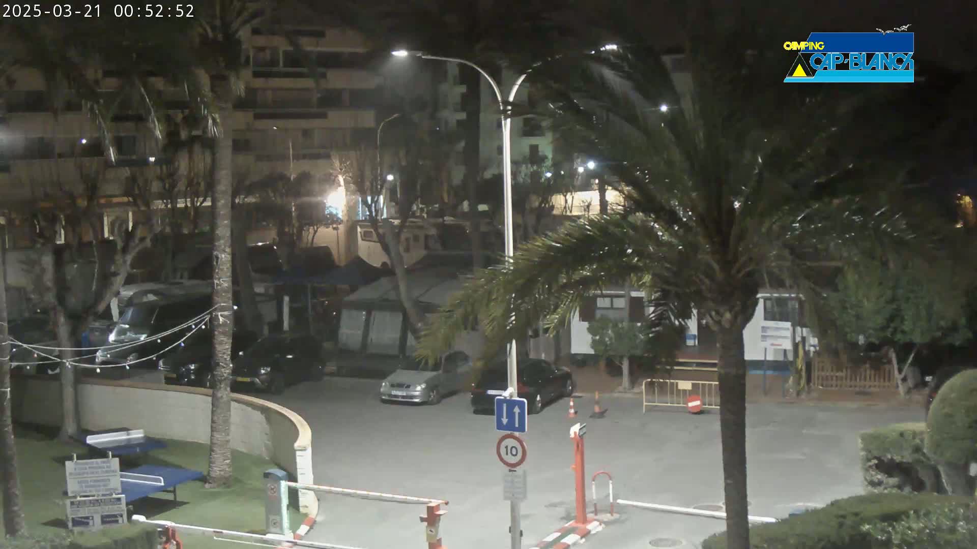 A nighttime view of a parking lot with cars and RVs, palm trees, and buildings under a dark, possibly windy sky.
