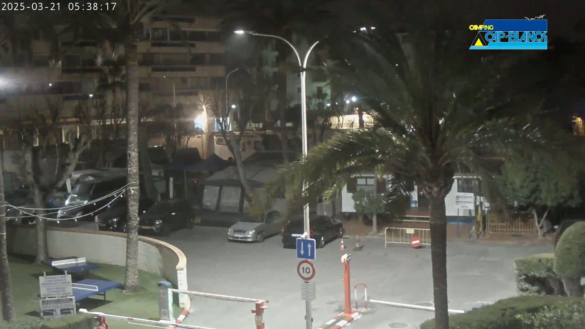 A nighttime view of a parking lot surrounded by palm trees and buildings, with several cars parked and a speed limit sign visible.