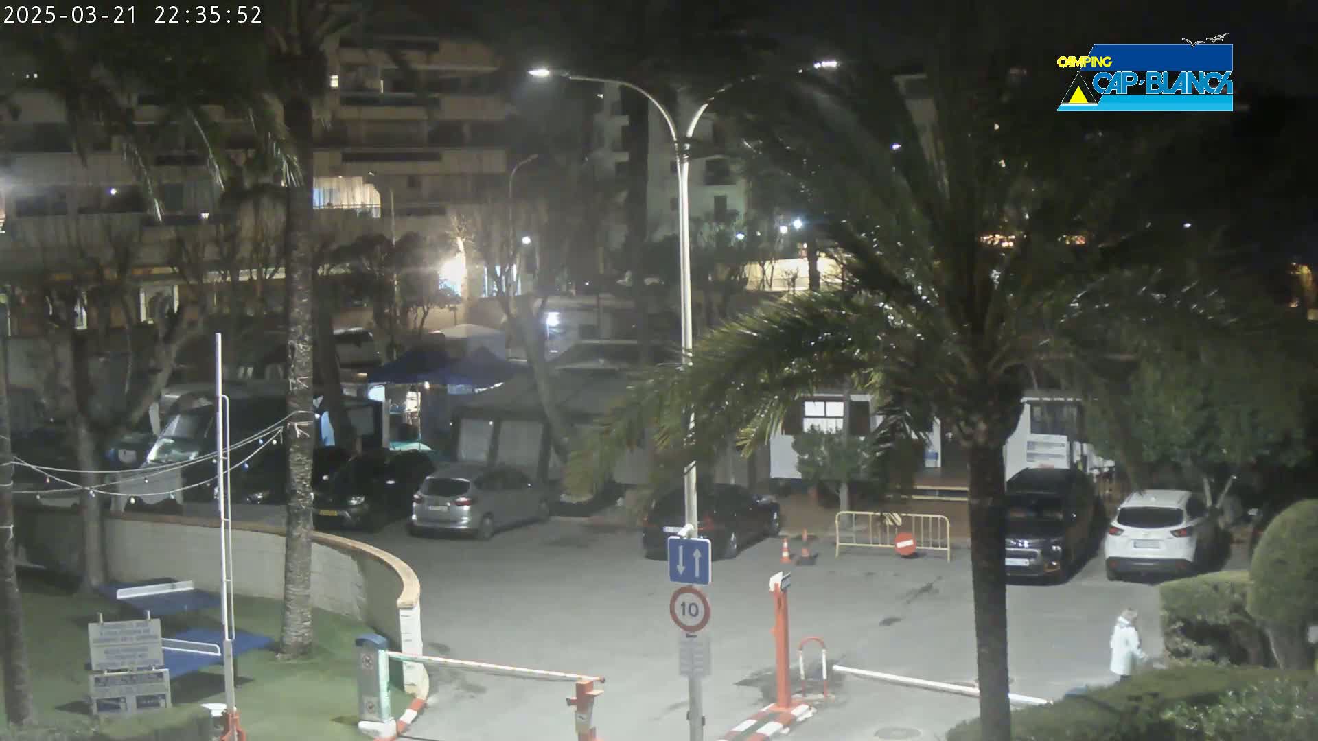 A nighttime scene of a parking lot with cars, palm trees, and buildings under a partly cloudy sky.