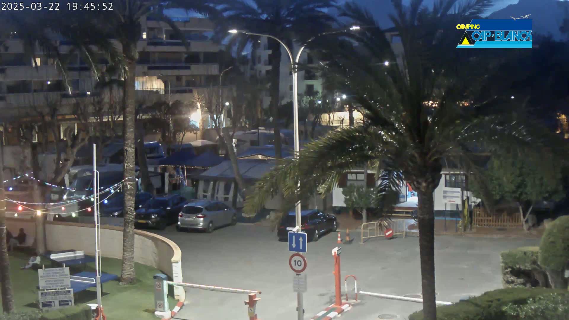 At night, under a dark sky, a parking lot with several cars is surrounded by palm trees, buildings, and tents, with strings of lights illuminating the scene.