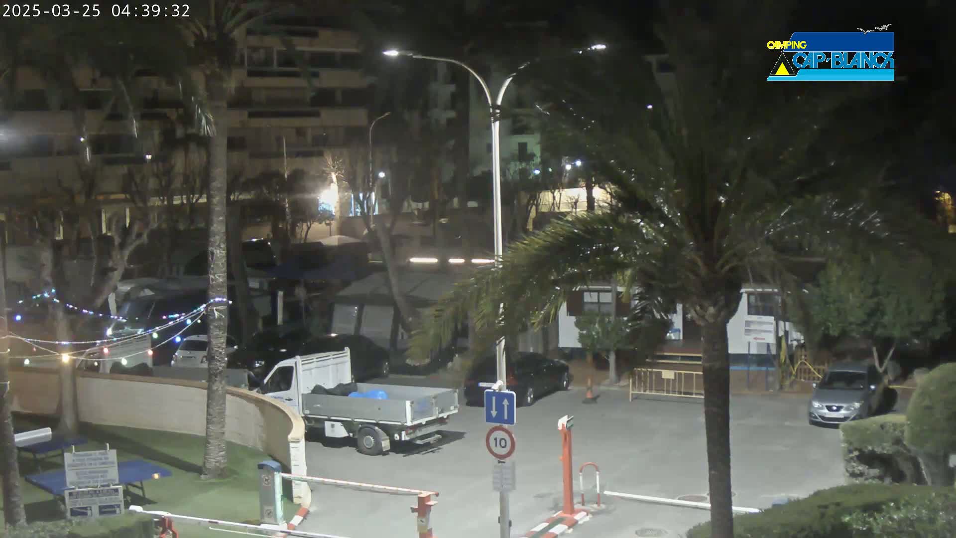 At night, a parking lot with several cars and a flatbed truck is illuminated by streetlights amongst palm trees and apartment buildings.