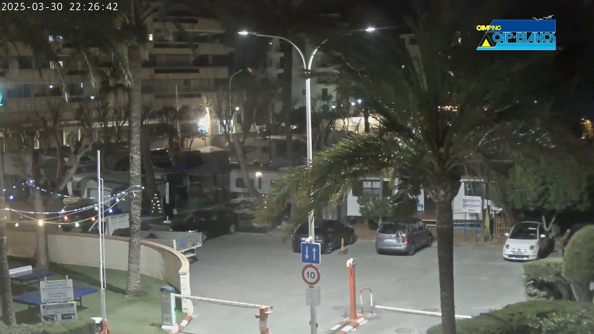 A nighttime parking lot with several cars and RVs, surrounded by palm trees and buildings, under a clear sky.