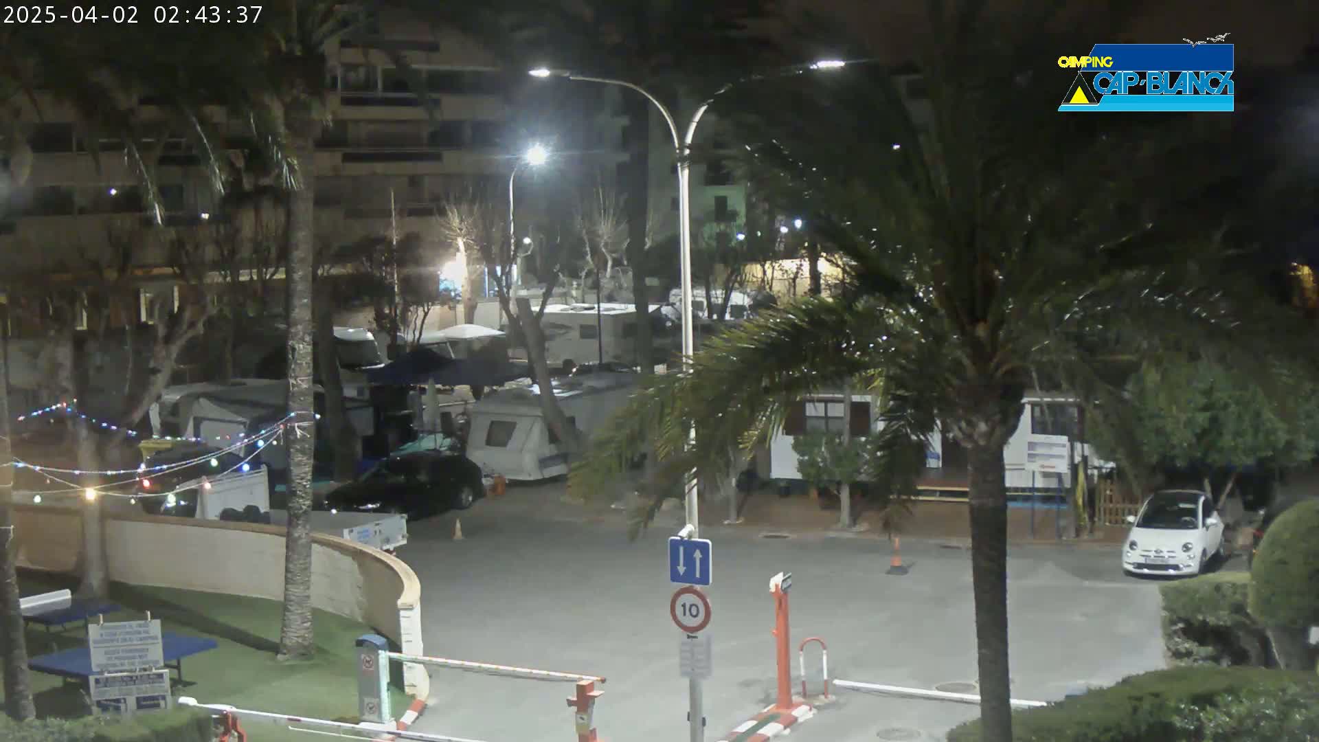 At night, a parking lot with several RVs and cars is illuminated by streetlights, surrounded by palm trees and buildings.