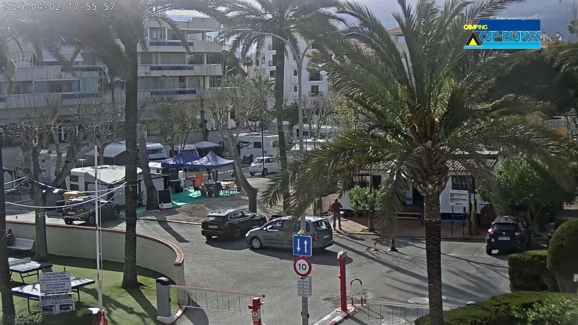 A sunny outdoor scene shows a parking lot with several cars and camper vans near a multi-story building and palm trees.