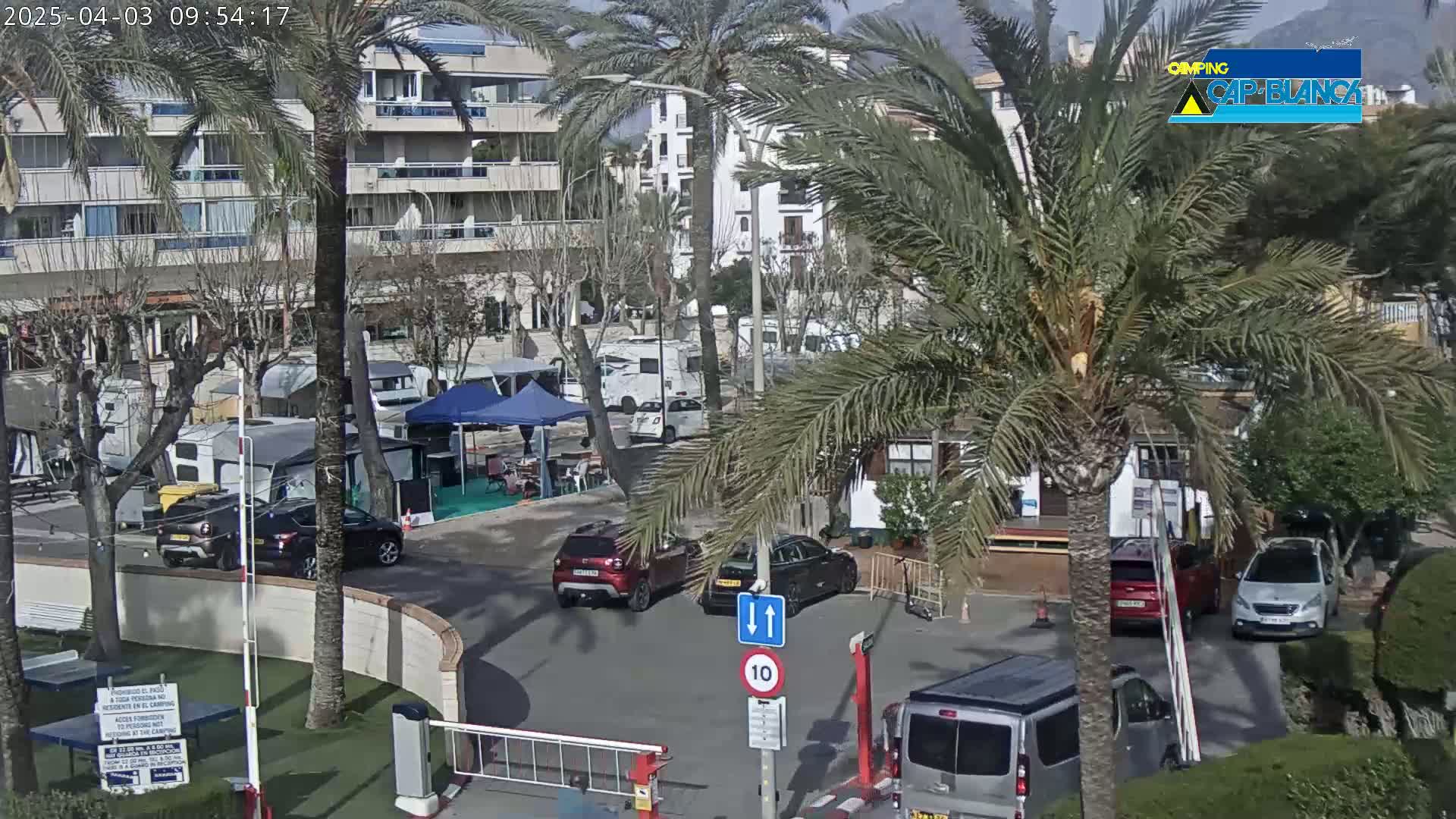 A sunny day at a campground with several cars and RVs parked near a building, palm trees, and a gated entrance.