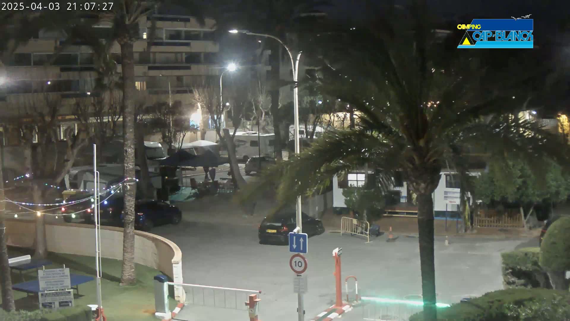 A nighttime view of a parking lot and campground with several cars and RVs, surrounded by palm trees and buildings under partly cloudy skies.