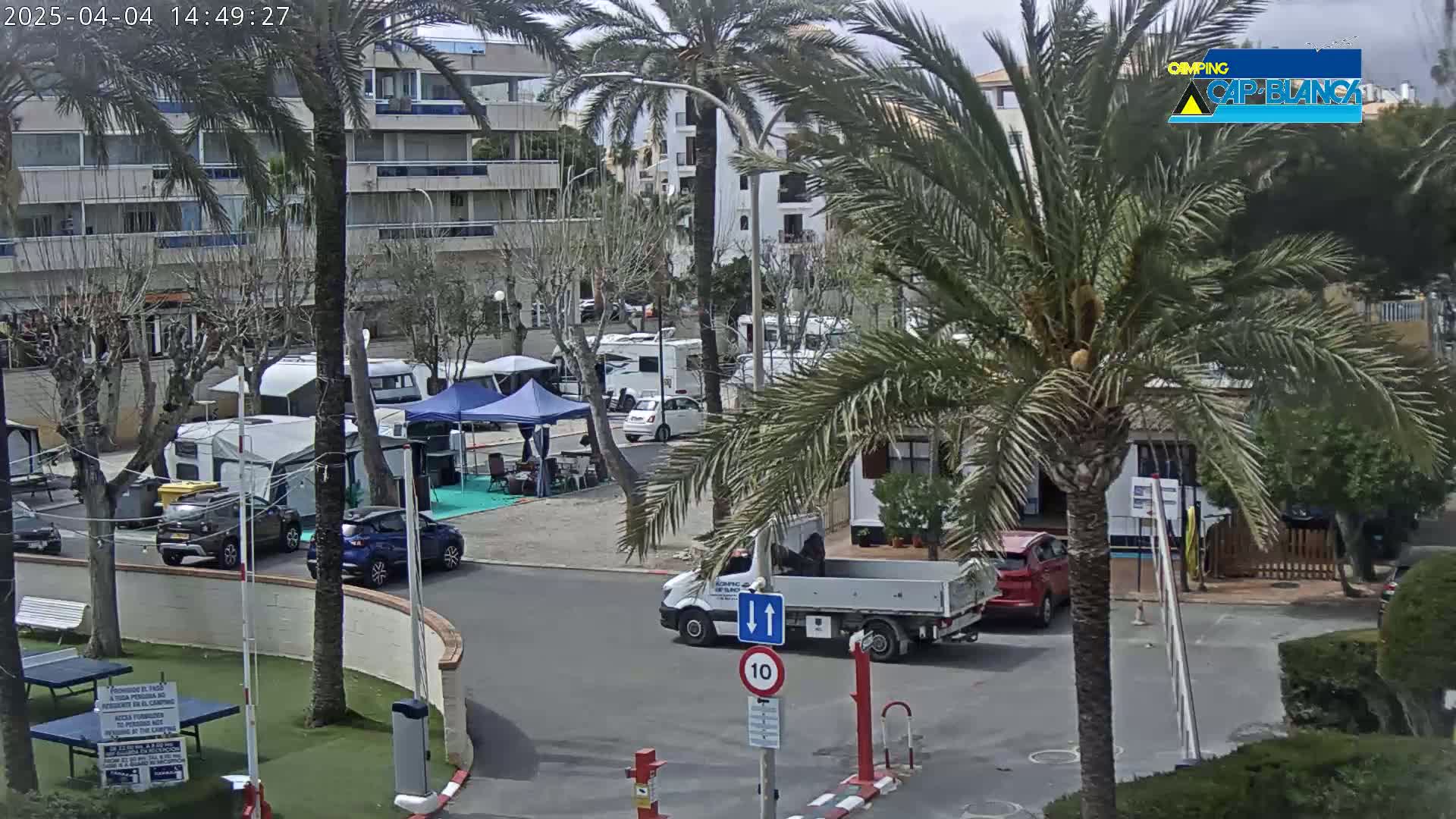 A parking lot with numerous recreational vehicles and cars, under a partly cloudy sky, is surrounded by palm trees and buildings.