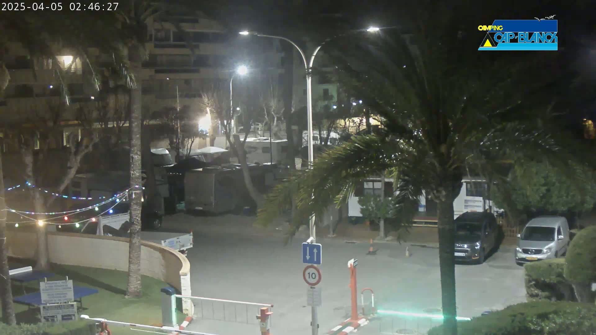 A nighttime view of a parking lot with several vehicles, palm trees, and buildings, illuminated by streetlights.