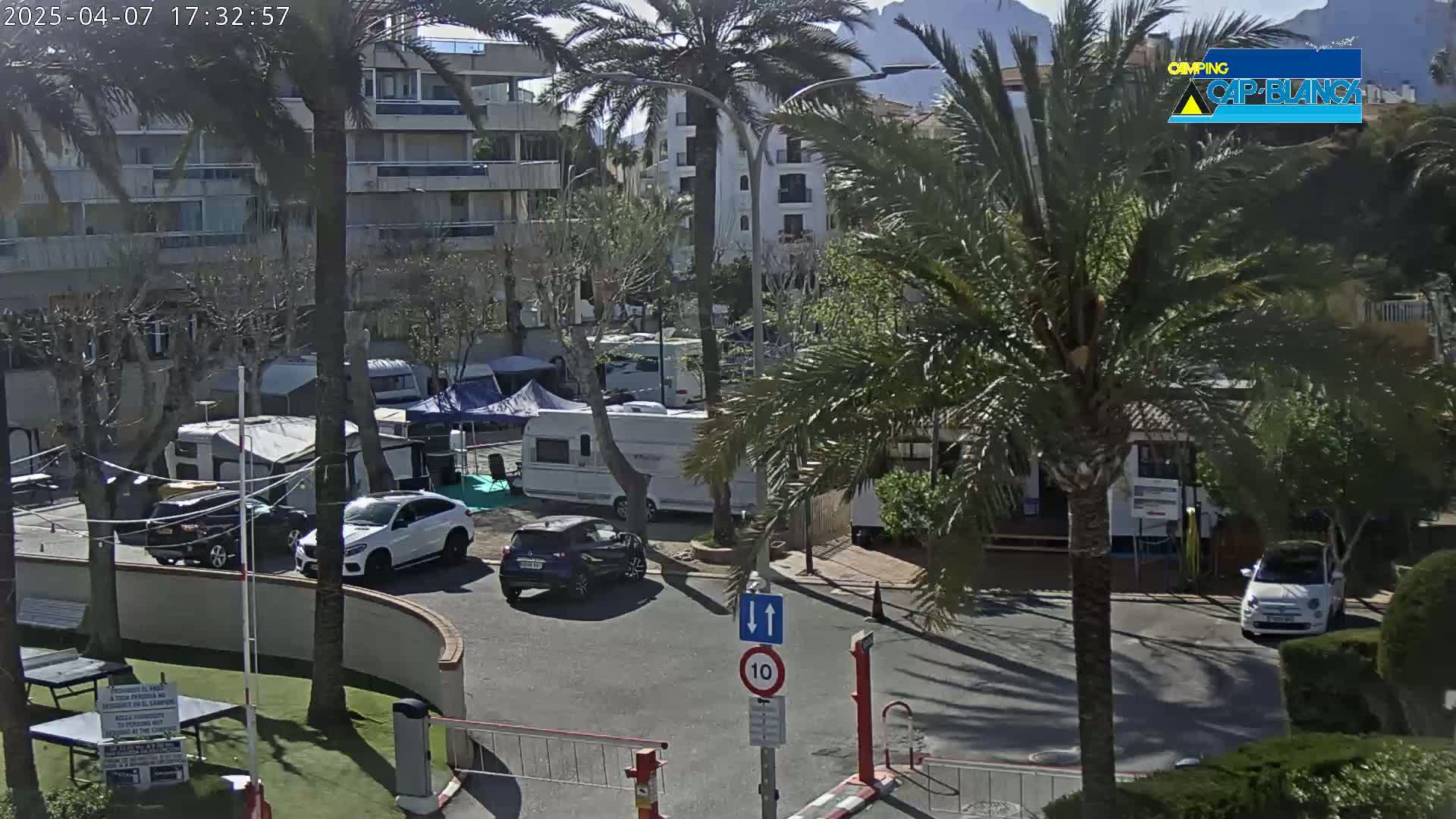 A sunny day at a campground with several RVs and cars parked near a multi-story building and palm trees.
