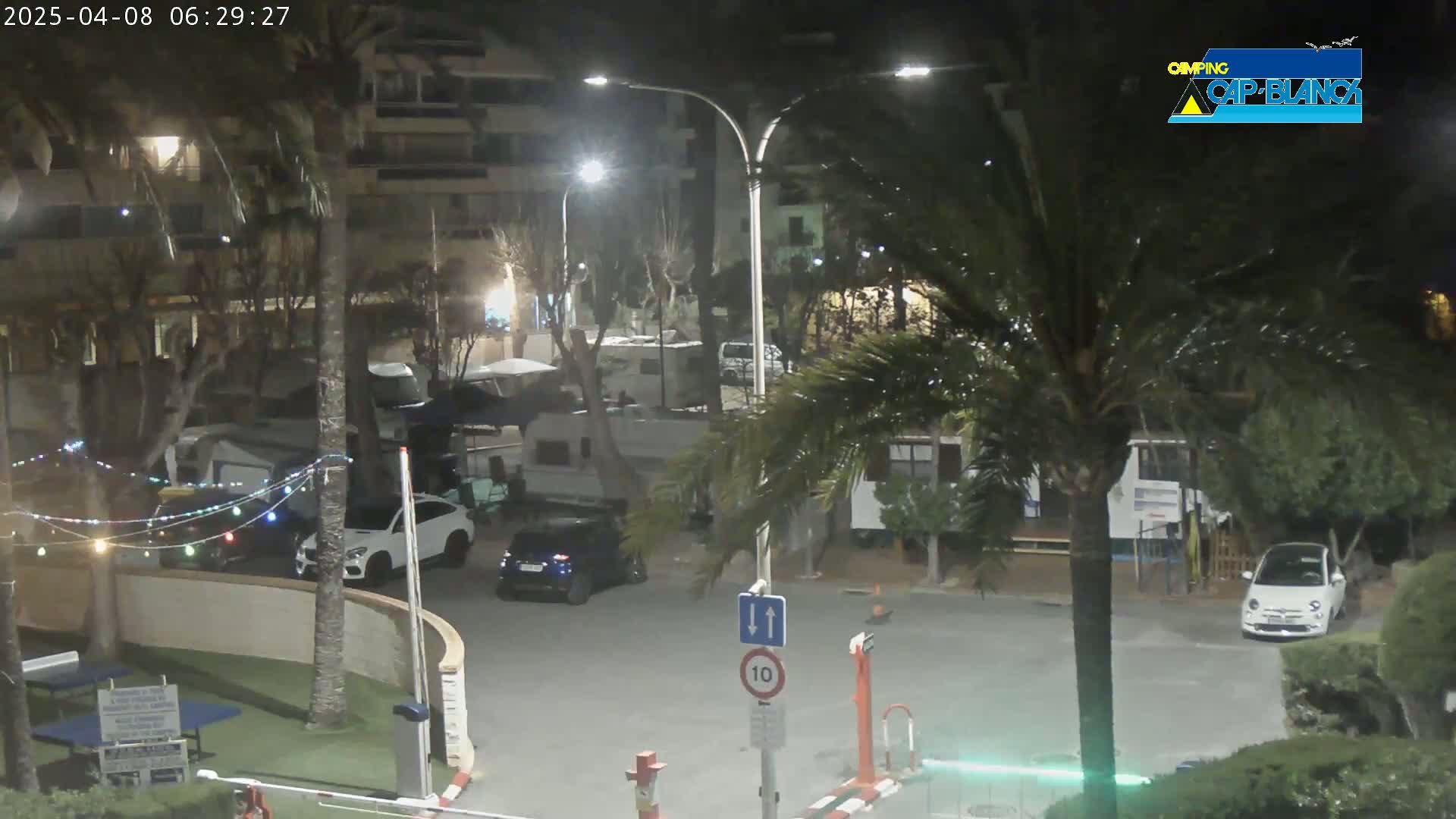 A nighttime, outdoor view of a parking lot with several cars and RVs, surrounded by trees and buildings, under artificial light.