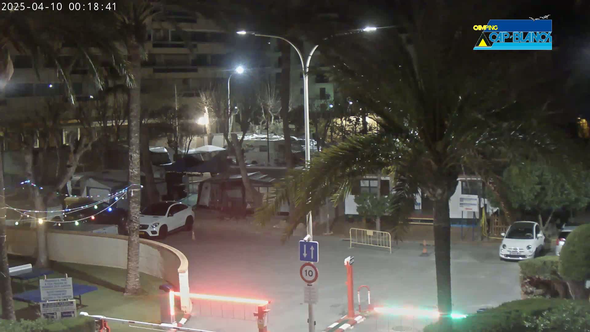 A nighttime view of a campground with cars parked, palm trees, and string lights illuminated.