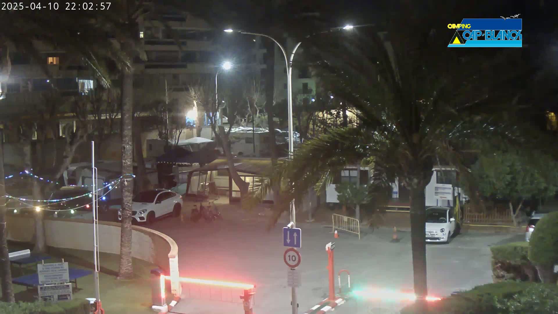 A nighttime view of a parking lot with cars, palm trees, and buildings, illuminated by streetlights.