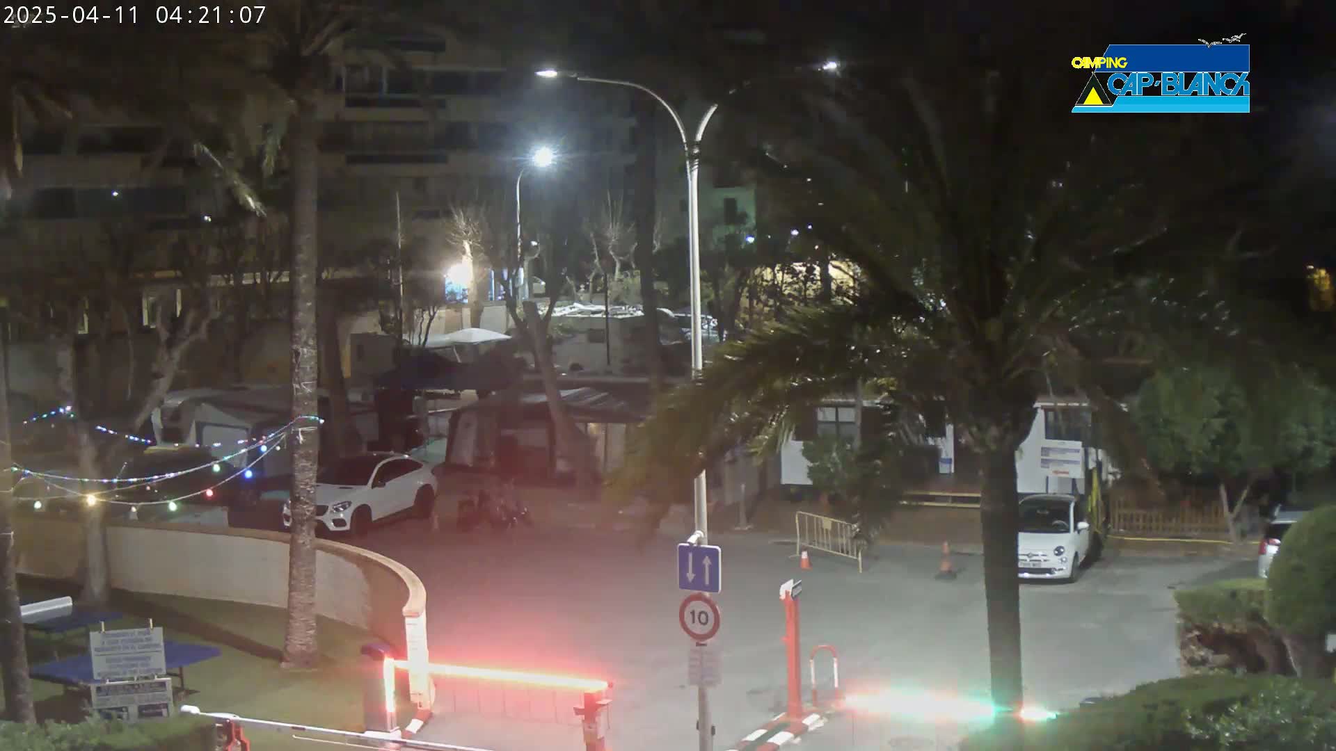 A nighttime scene of a campsite with cars parked near buildings and palm trees, under windy conditions.
