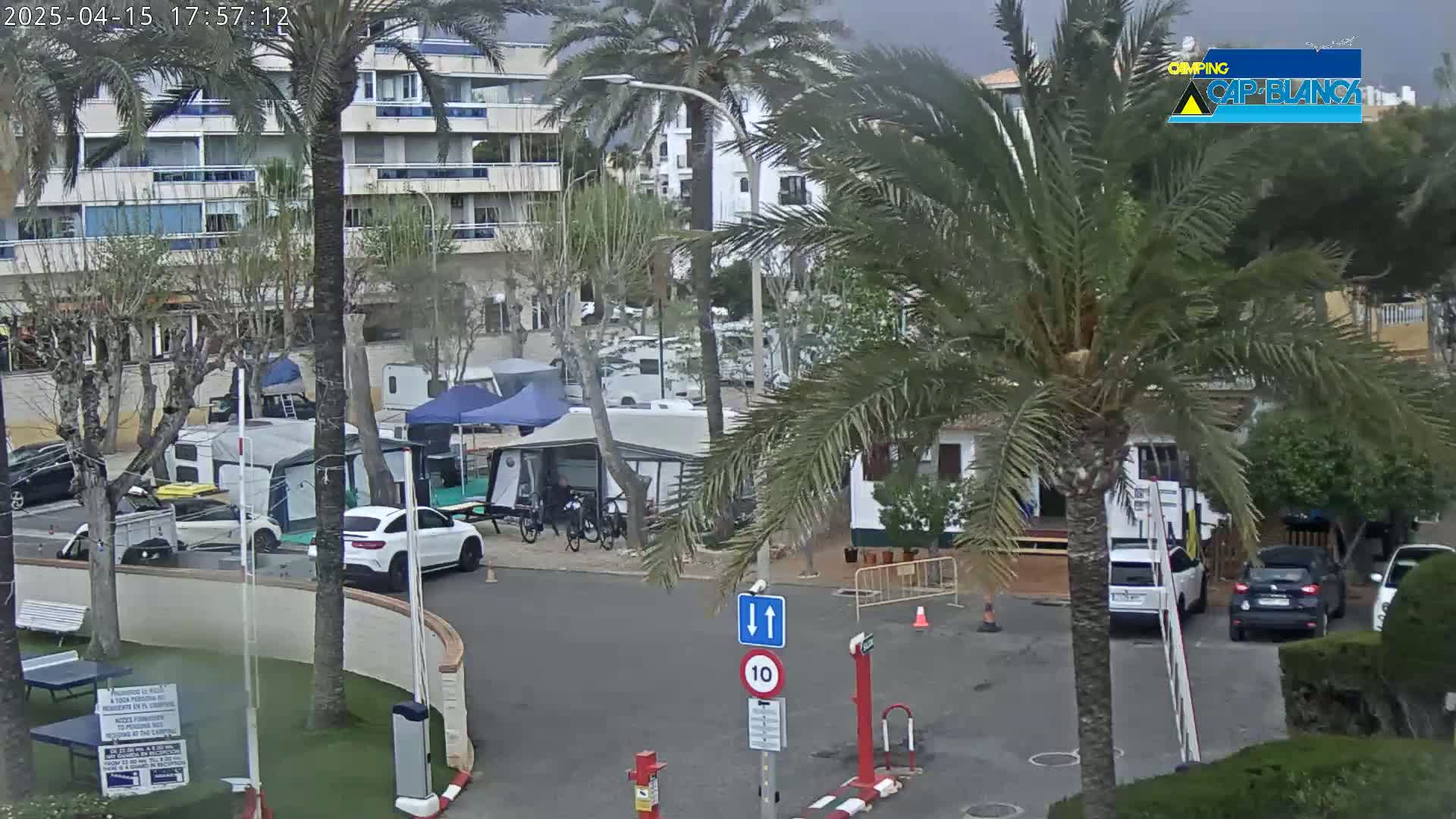 A mostly cloudy day shows a parking lot with several cars, RVs, and tents situated near apartment buildings and palm trees.