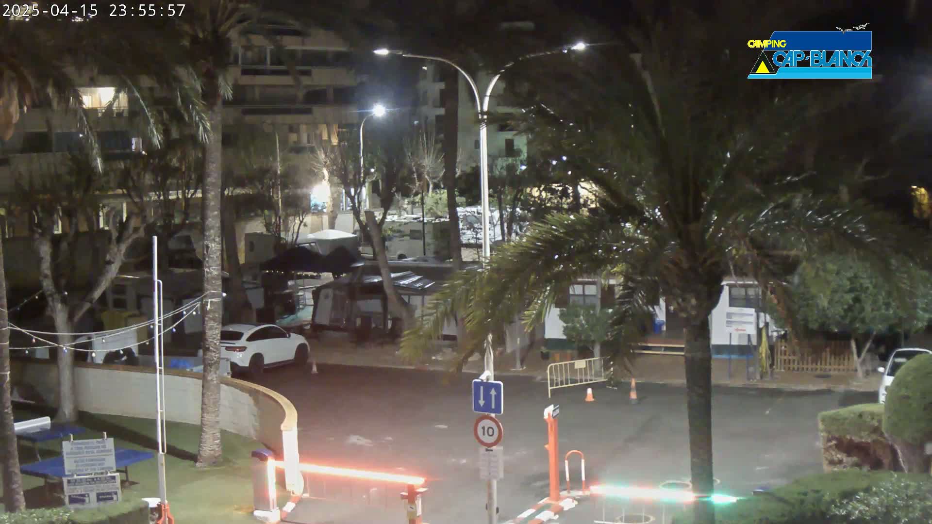 A nighttime view of a parking lot with palm trees, buildings, and a few cars, illuminated by streetlights.