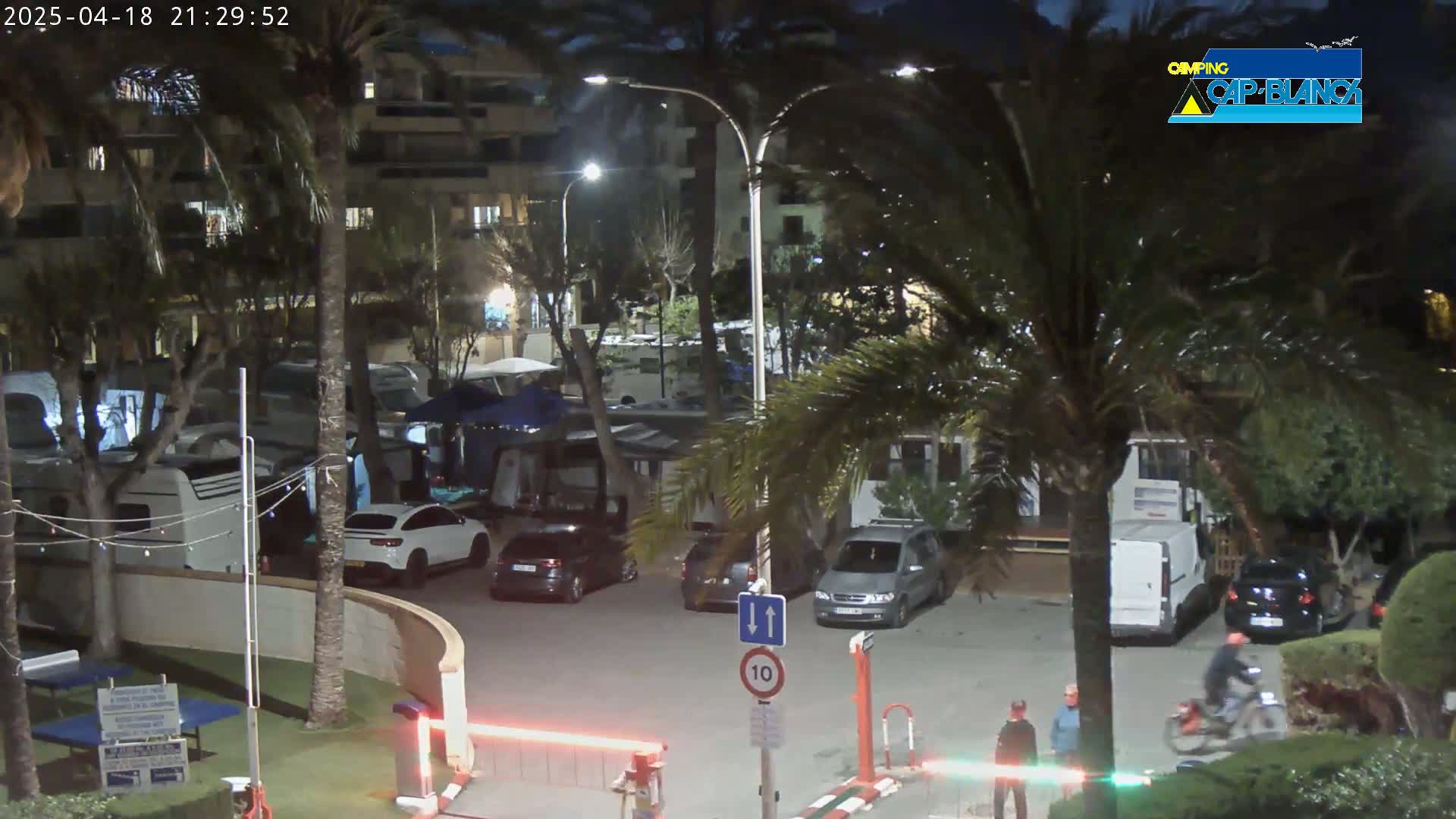 A nighttime scene in a parking lot surrounded by palm trees and buildings, with several cars, RVs, and a person on a motorcycle.