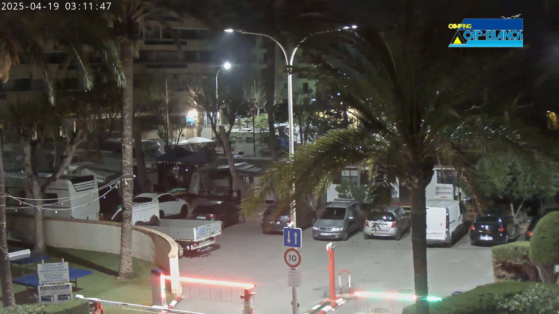 A nighttime view of a parking lot with several cars, RVs, and palm trees under artificial light.