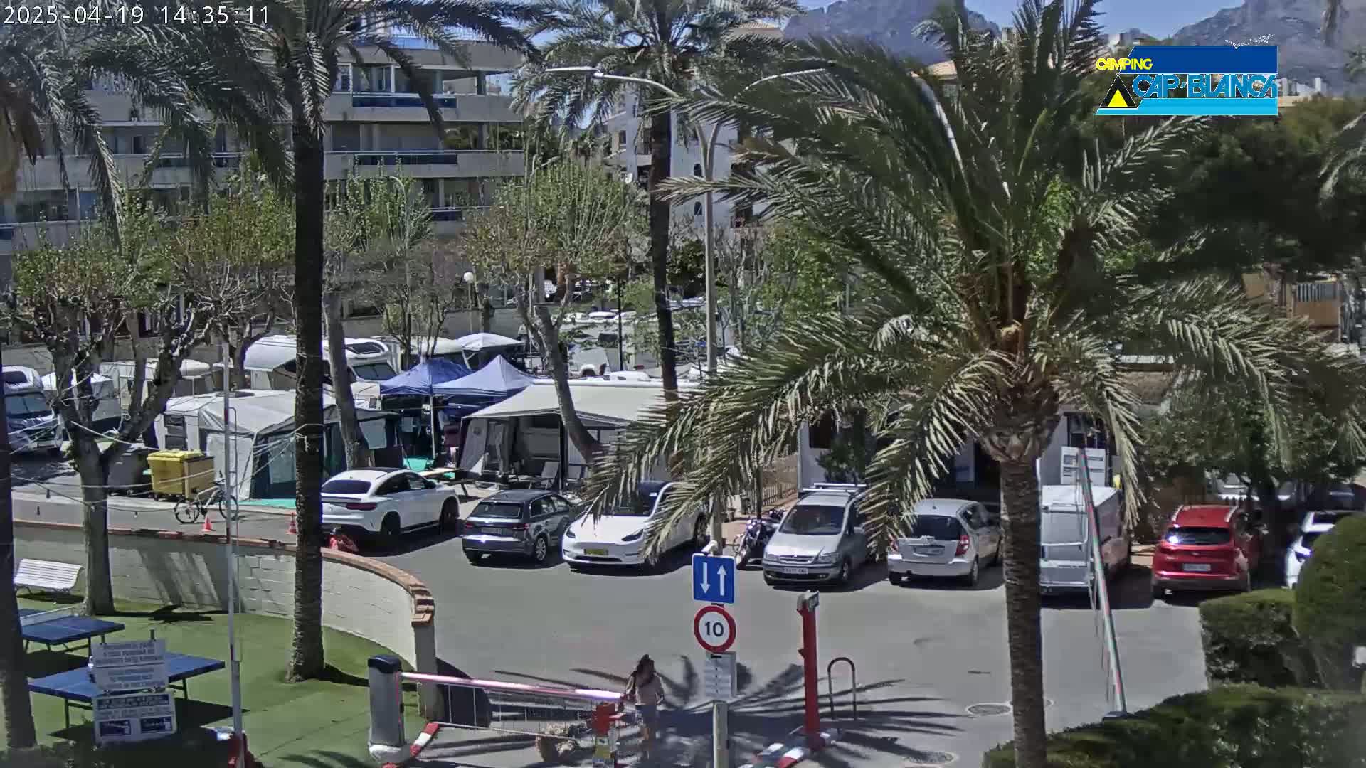 A sunny outdoor scene shows a parking lot with cars and RVs near palm trees and a multi-story building, under a mostly clear sky.