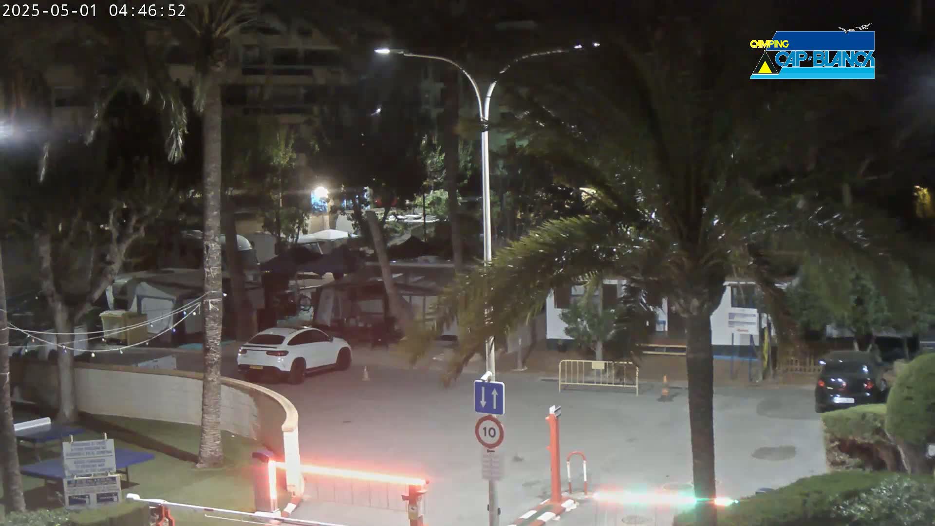A nighttime view of a parking lot with cars and RVs, surrounded by palm trees under partly cloudy skies.